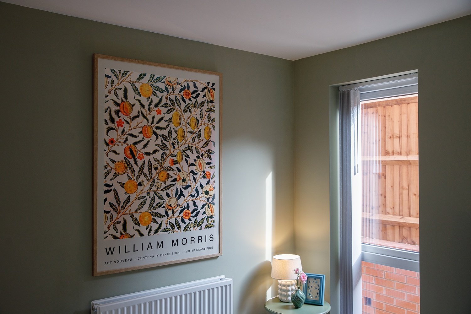 Interior of a room with a framed art print on the wall, a small table with a lamp, a pink flower, and a blue picture frame, and an open window showing a wooden fence outside.