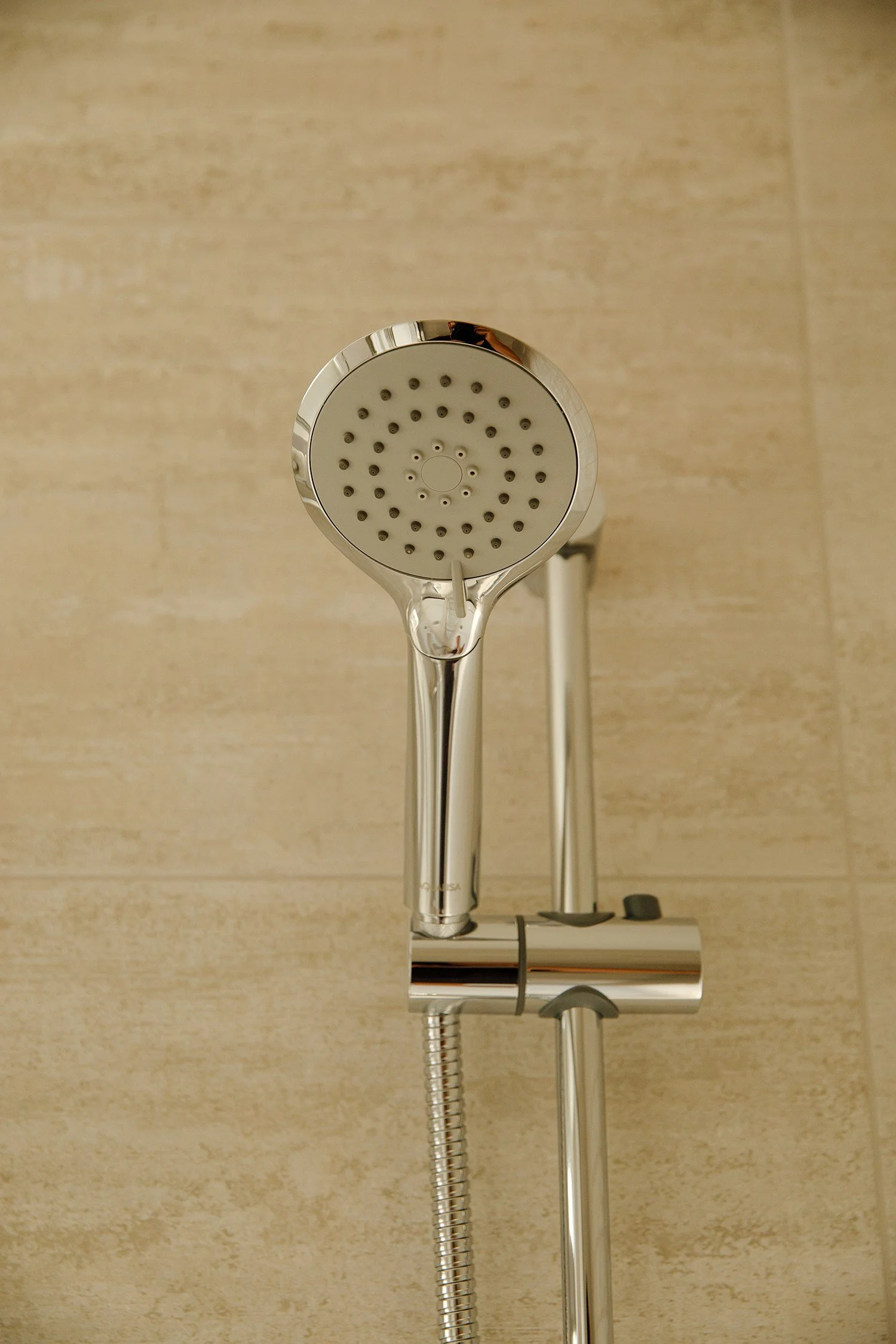A showerhead mounted on a wall with beige tiles, connected to a flexible hose and a bathroom fixture.