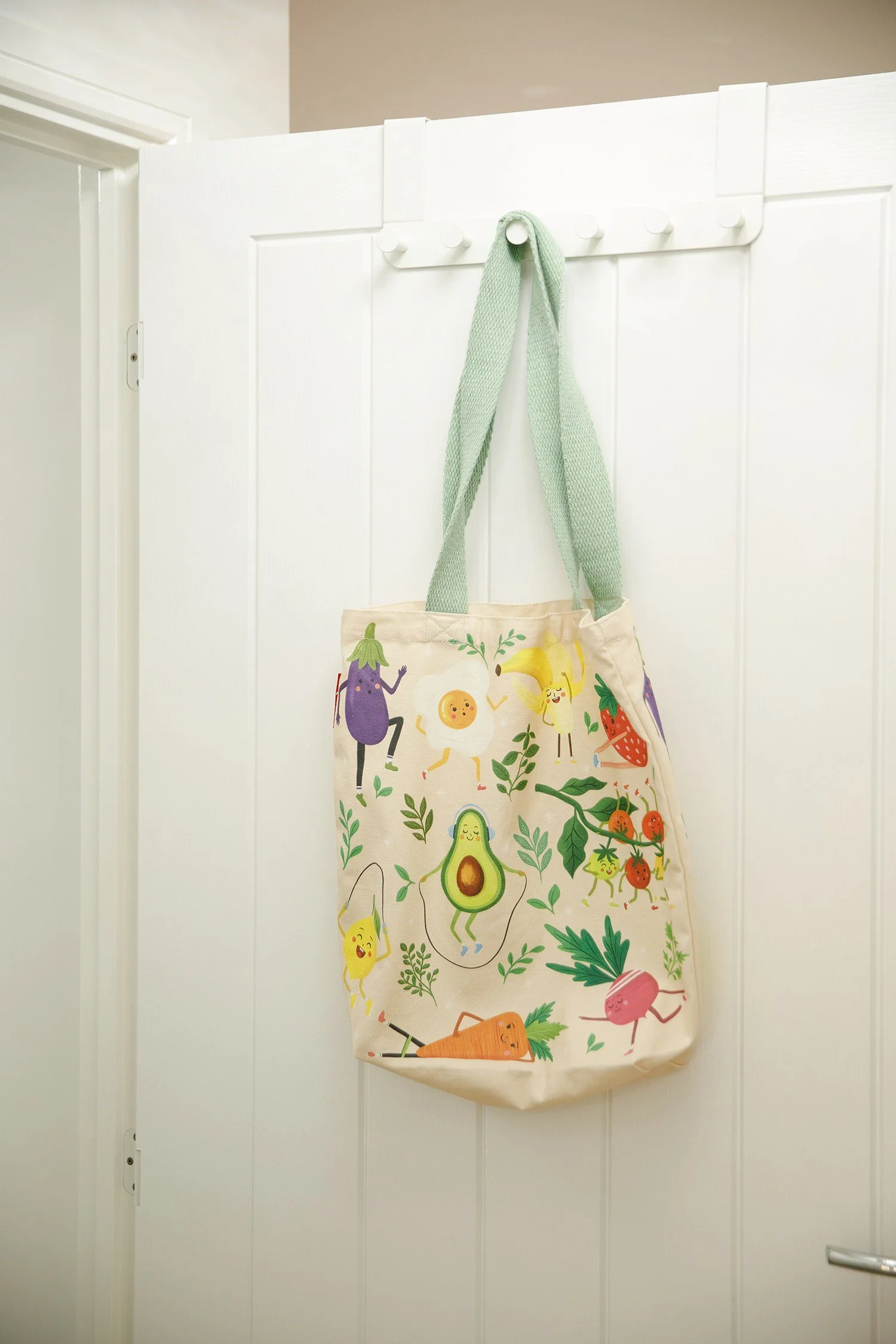 A tote bag hanging on a white wall hook with green straps. The bag features colorful cartoon vegetables with smiling faces and various greenery.