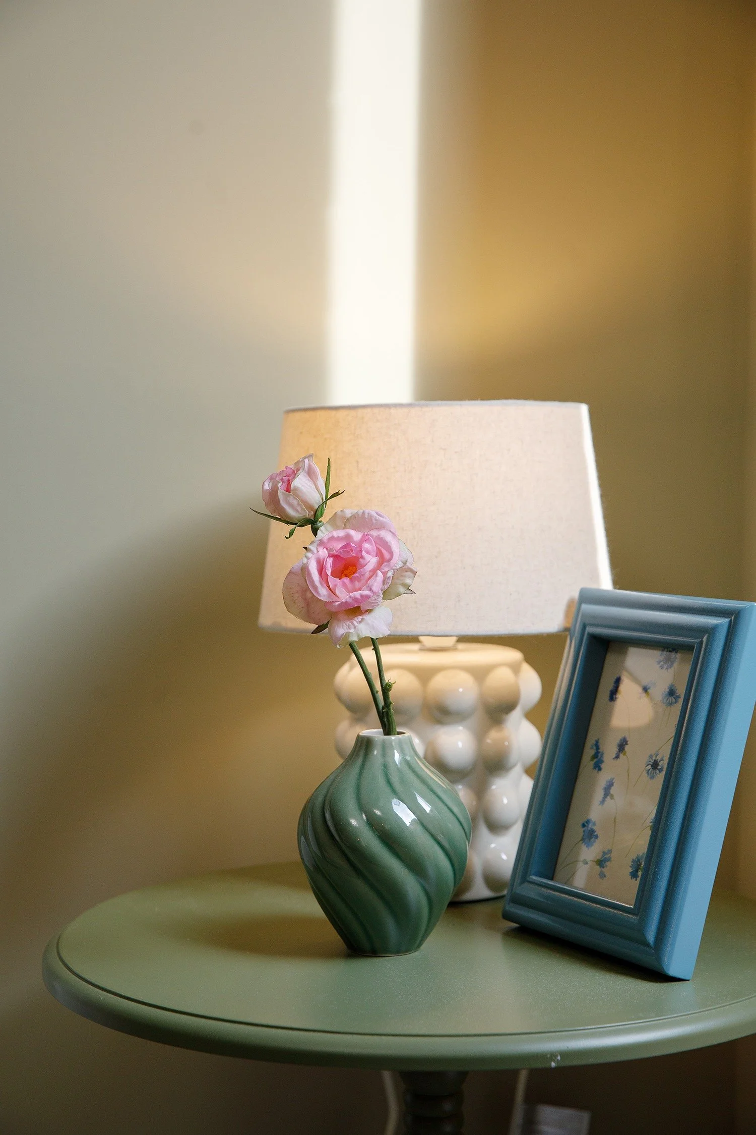 A green vase with pink flowers on a table, beside a framed picture, and a white lamp in the background.