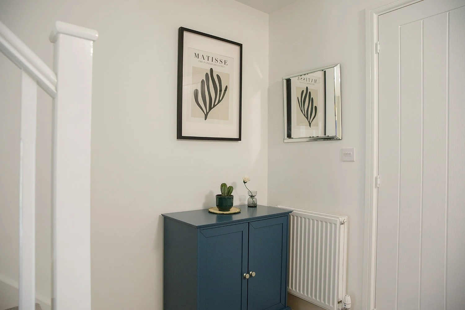 Interior of a room with white walls, a blue cabinet with two doors, a small cactus and a flower in a glass on top, framed artwork of a black abstract plant and a mirror on the wall, a white radiator, and a white door.