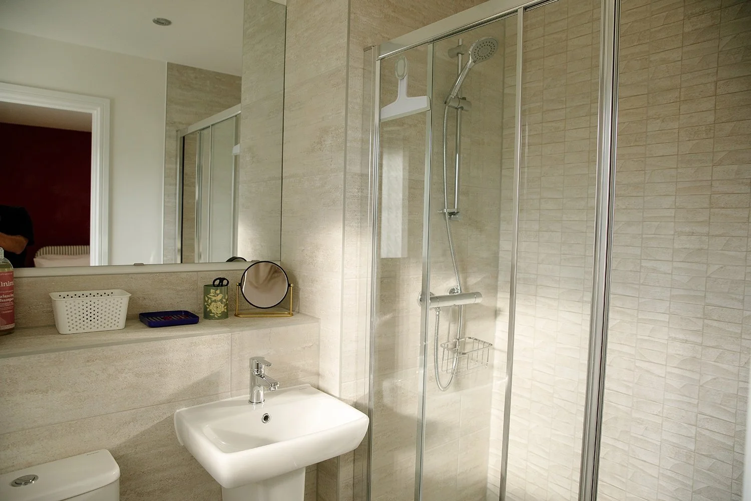 Bathroom with beige tiled walls, a glass shower enclosure, white sink, mirror, and various bathroom items on the counter.