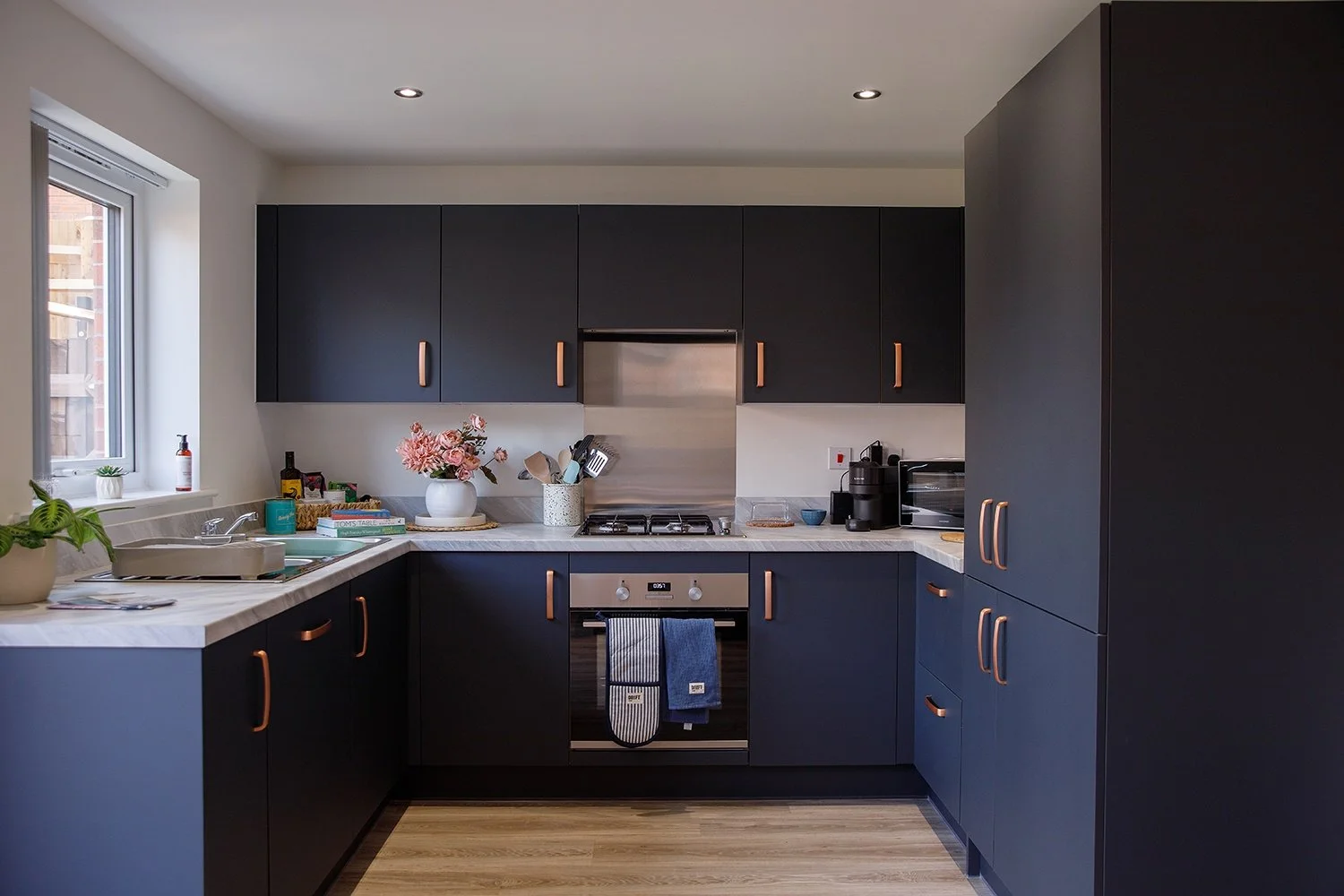Modern kitchen with black cabinets, white marble countertops, and wooden flooring. Includes sink, stove, oven, coffee machine, microwave, and decorative plants, with a window on the left.