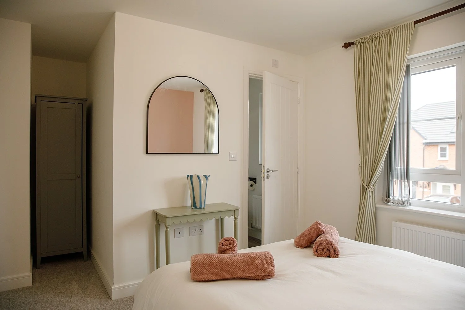 A bedroom with a white bed, pink towels on the bed, a window with beige and white striped curtains, a mirror on the wall, a small table near the door with a decorative vase, and a wardrobe in the corner.