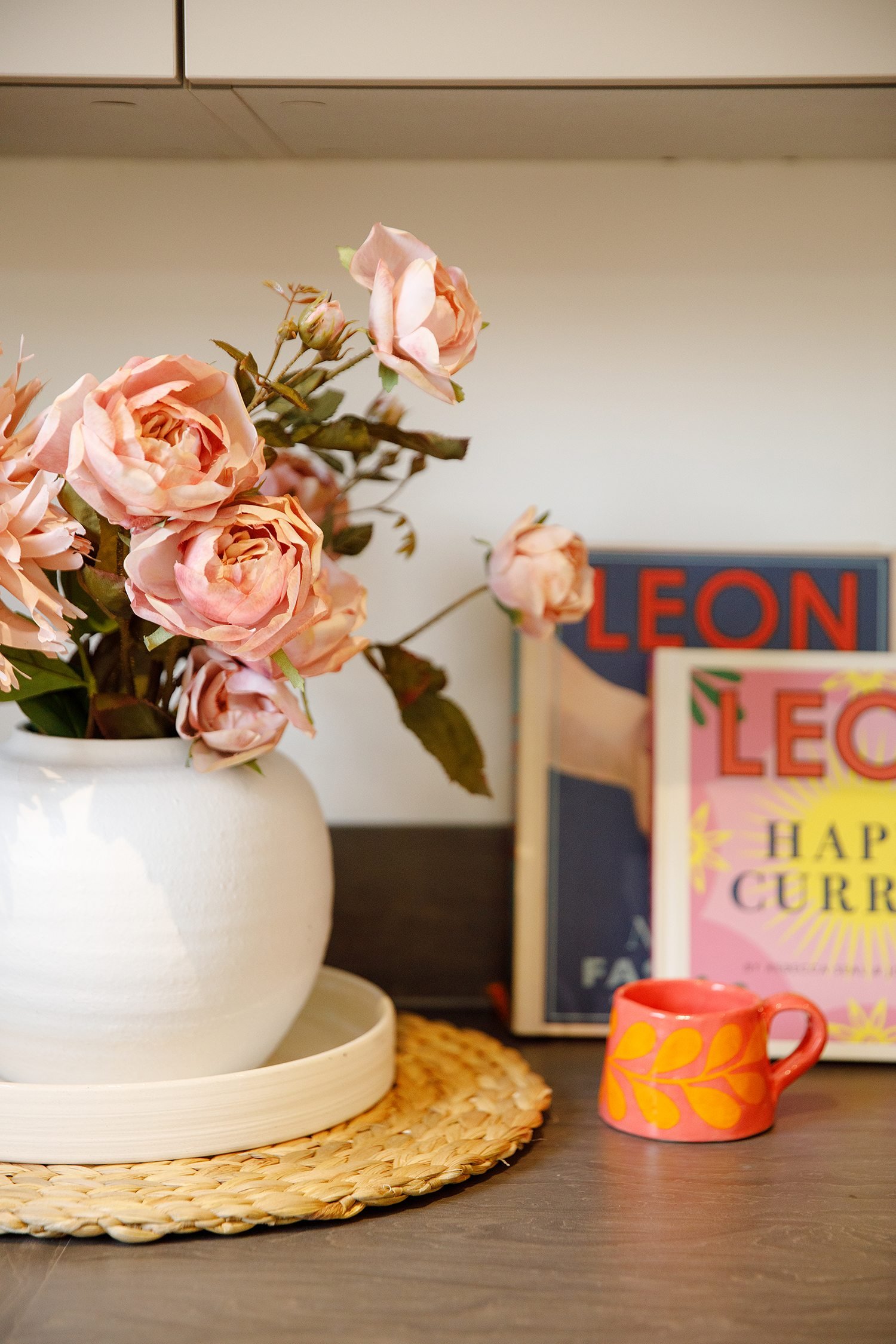 A white vase with pinkish-peach artificial roses on a woven placemat. To the right, a colorful mug with yellow leaves and pink background, and behind it, colorful books including one titled 'LEON'. The scene appears to be on a wooden table against a 
