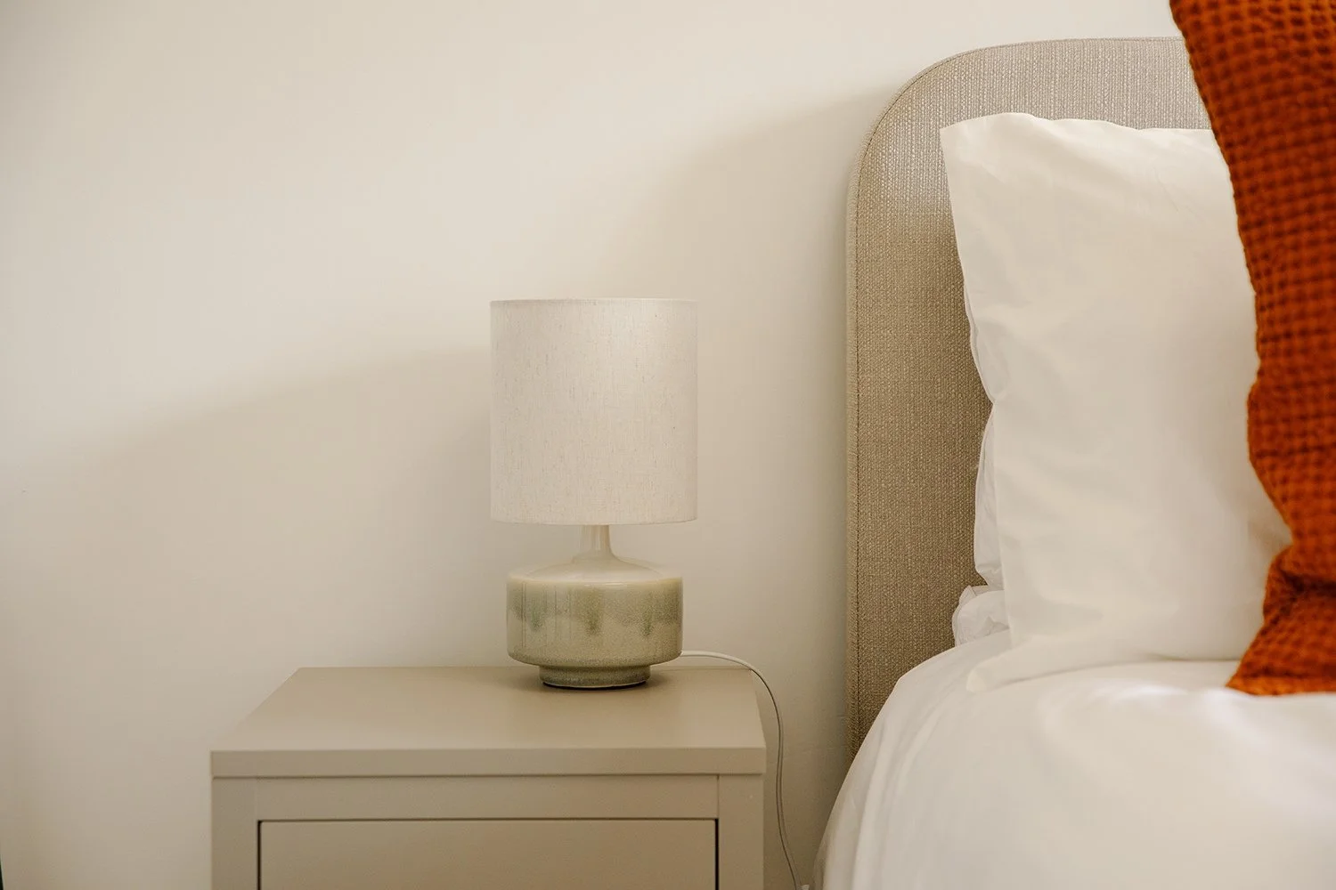 A bedside corner with a beige headboard, white pillow, orange pillow, beige nightstand, and a table lamp with a white shade and greenish base against a cream-colored wall.