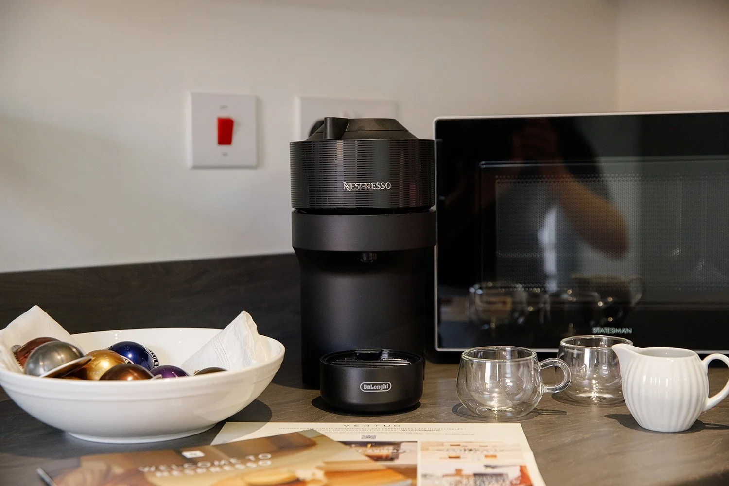 Coffee station with a Nespresso machine, a DeLonghi milk frother, two glass cups, a small white pitcher, a bowl of coffee capsules, and some papers on a dark wooden counter.