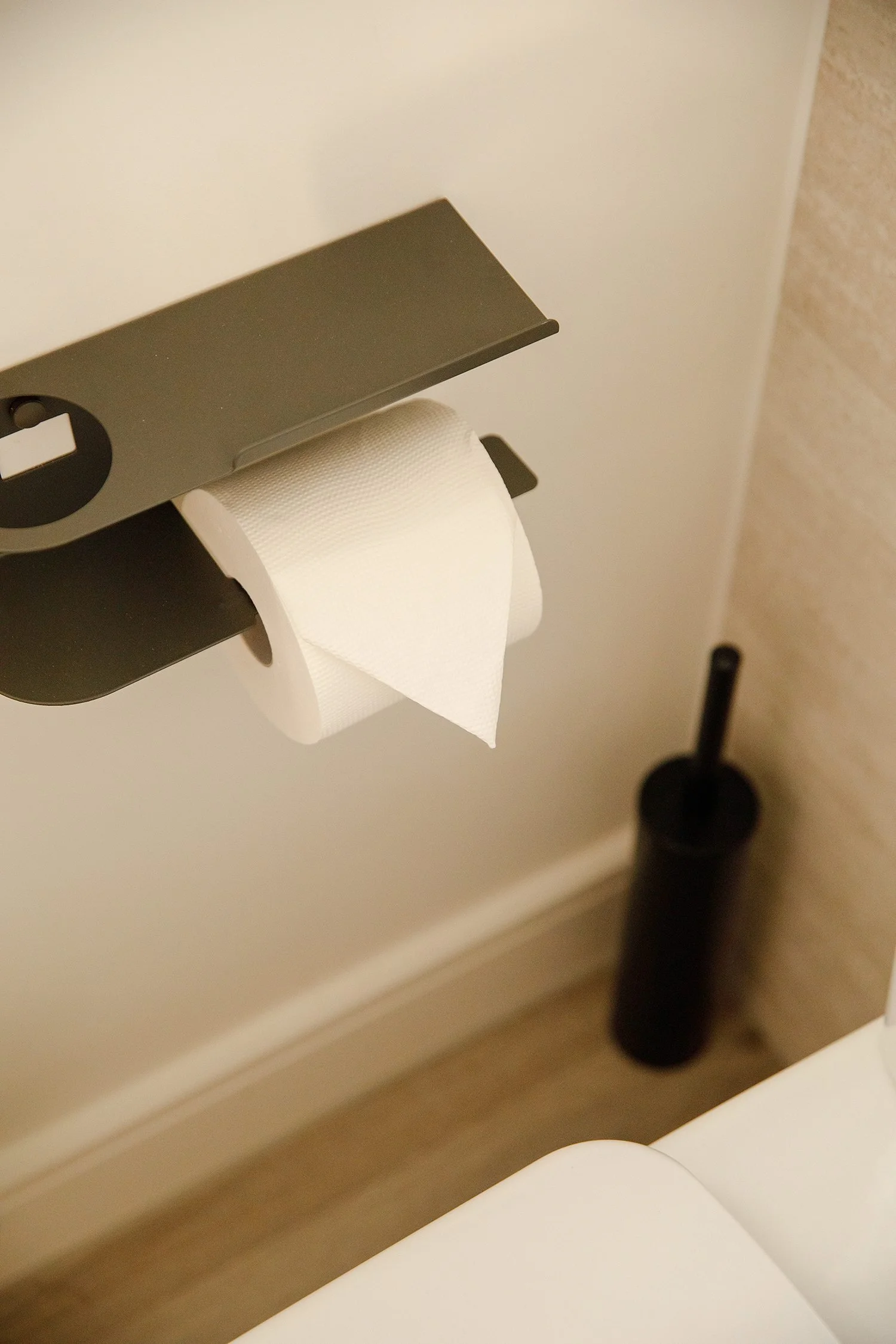 A roll of toilet paper hung on a metal bathroom toilet paper holder, with a toilet brush visible nearby. The background includes parts of a white wall and a beige wall.