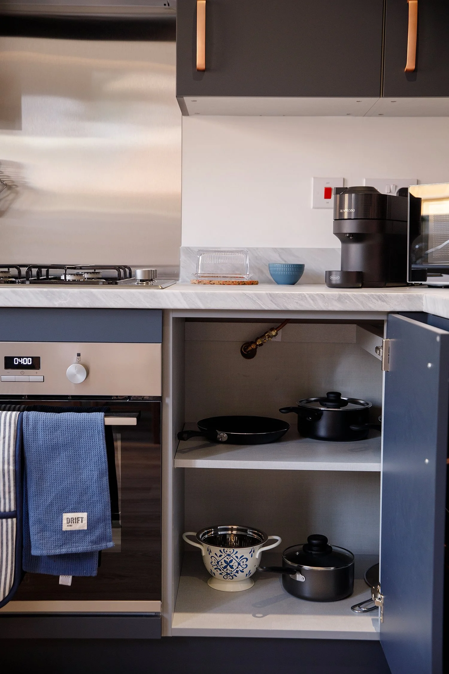 Kitchen cabinet with open lower section containing various cookware, including a patterned ceramic mixing bowl, a black skillet, and a small black pot, with a white marble countertop above.