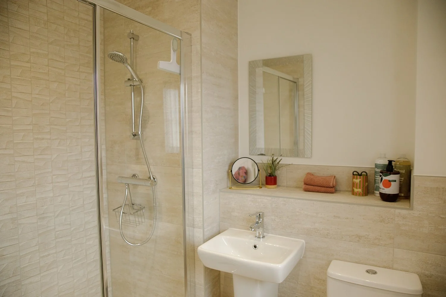 A modern bathroom with a walk-in shower, small sink, mirror, and beige tiled walls. There are toiletries and a small decorative plant on a shelf.