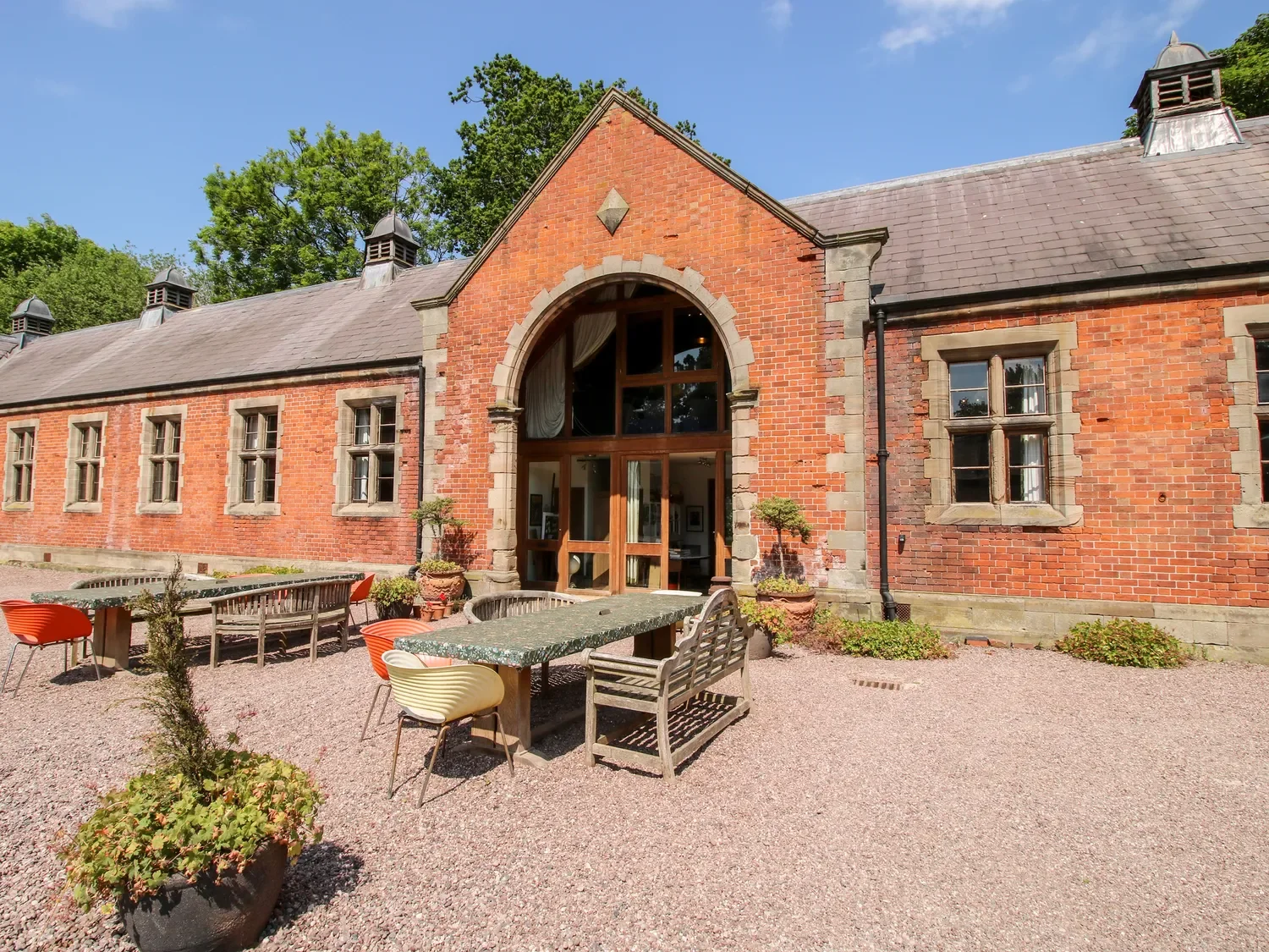 Exterior of a historic brick building with large arched and rectangular windows, a slate roof, and a small outdoor seating area with colorful chairs and potted plants in front.