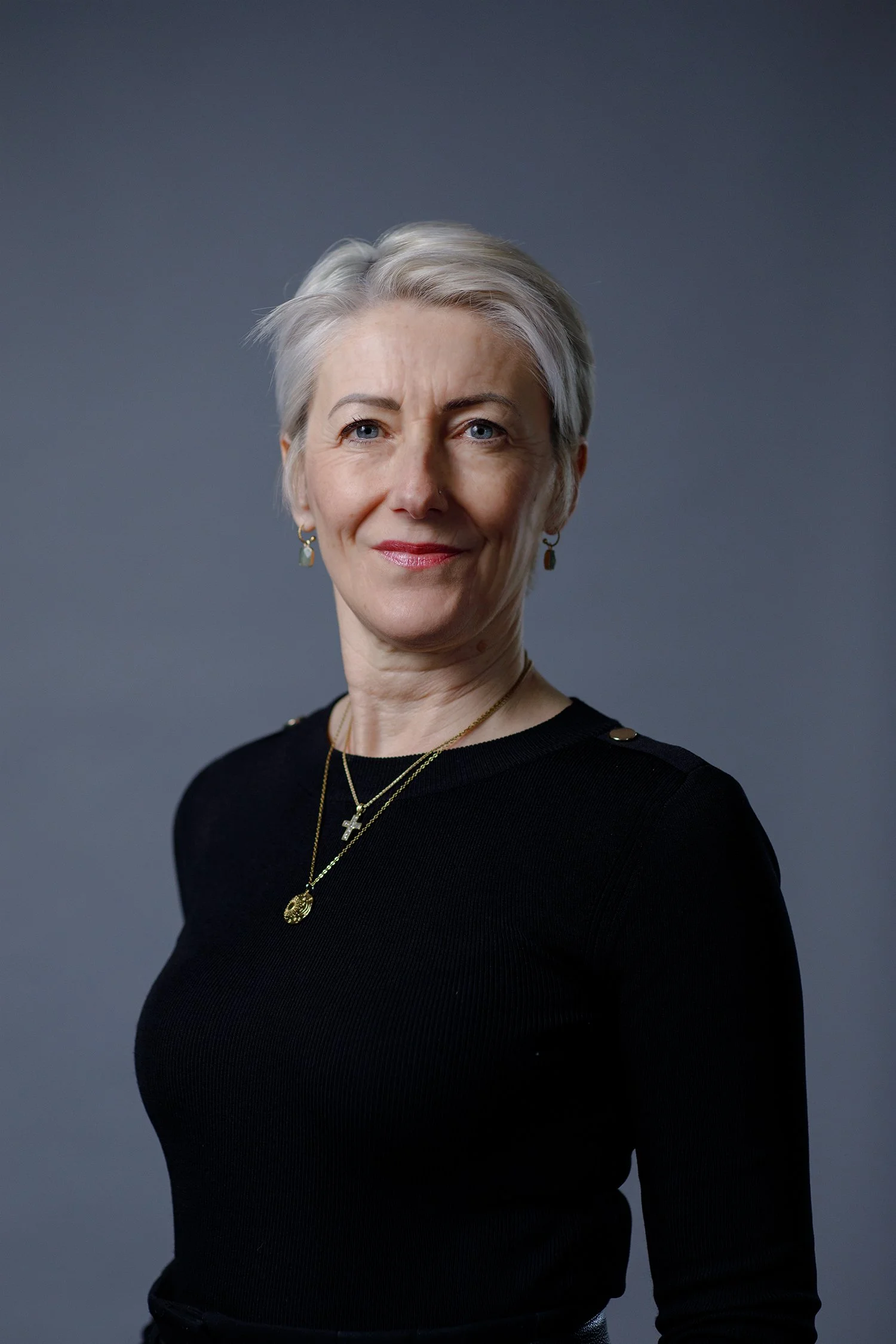 Portrait of Claire Wyon, wearing a black top and gold jewellery, standing against a dark blue background.