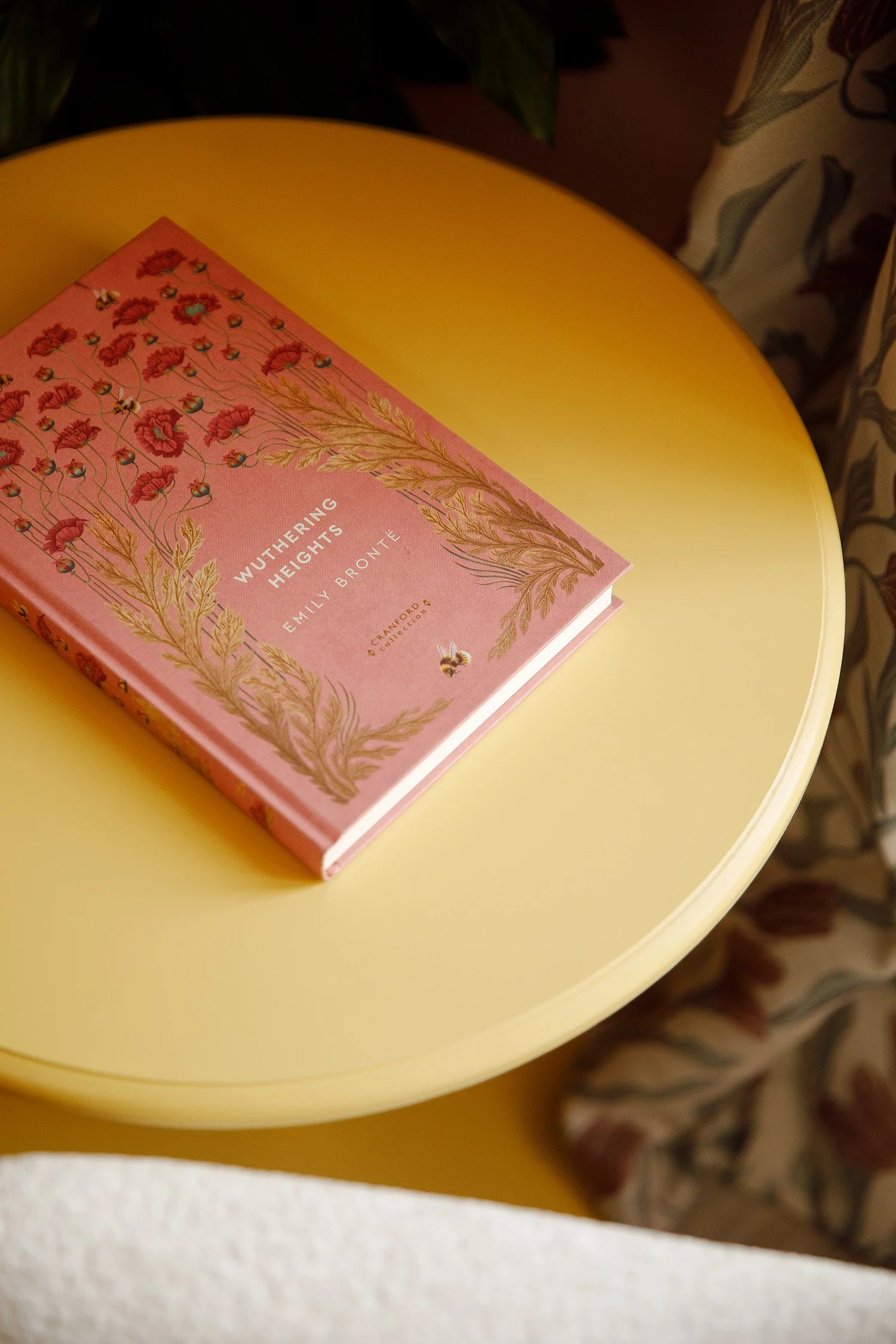 Pink book titled 'Wuthering Heights' by Emily Bronte resting on a yellow round table with a floral-patterned chair in the background.