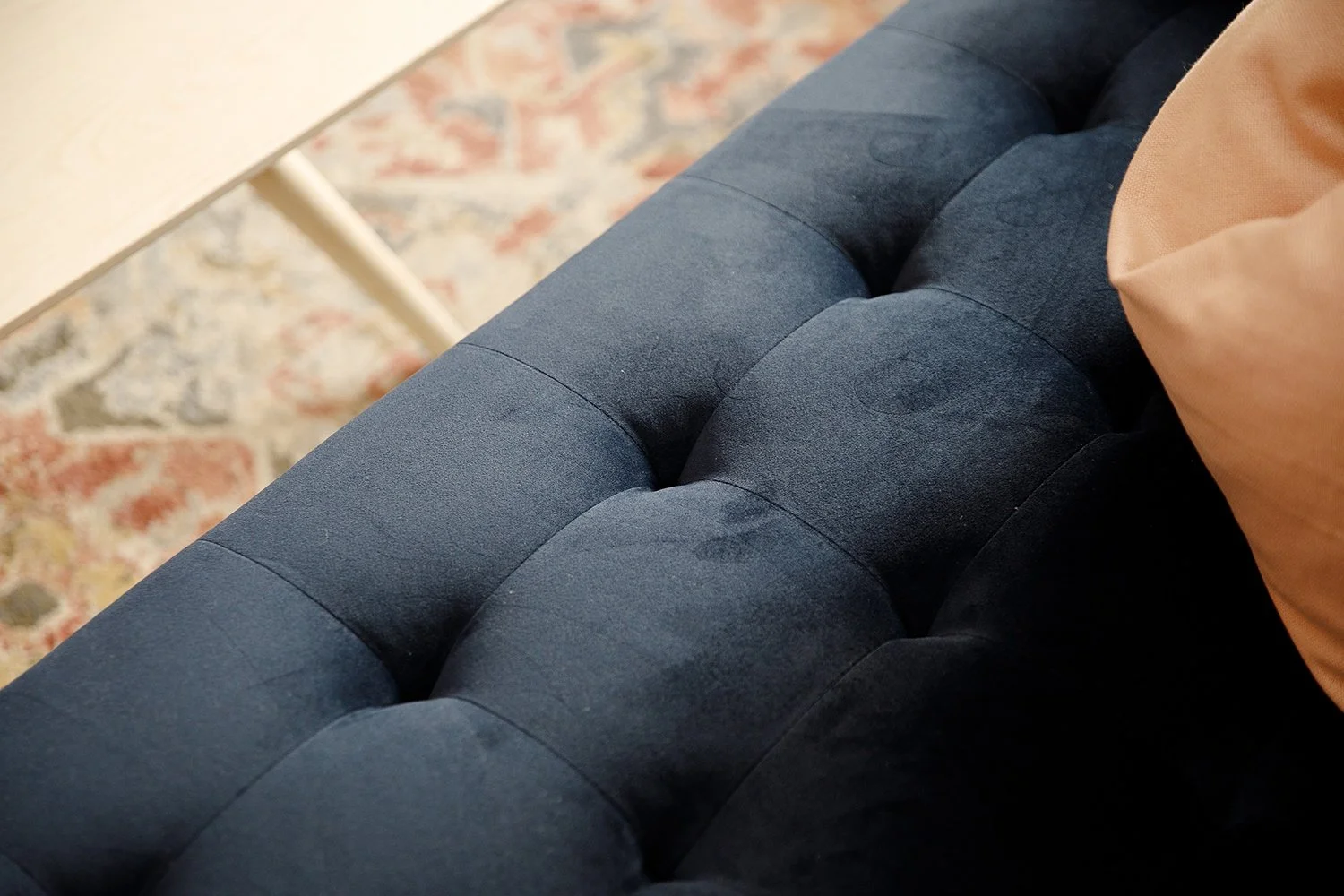 Close-up of a dark blue tufted sofa with a tan cushion in a room with a patterned area rug.