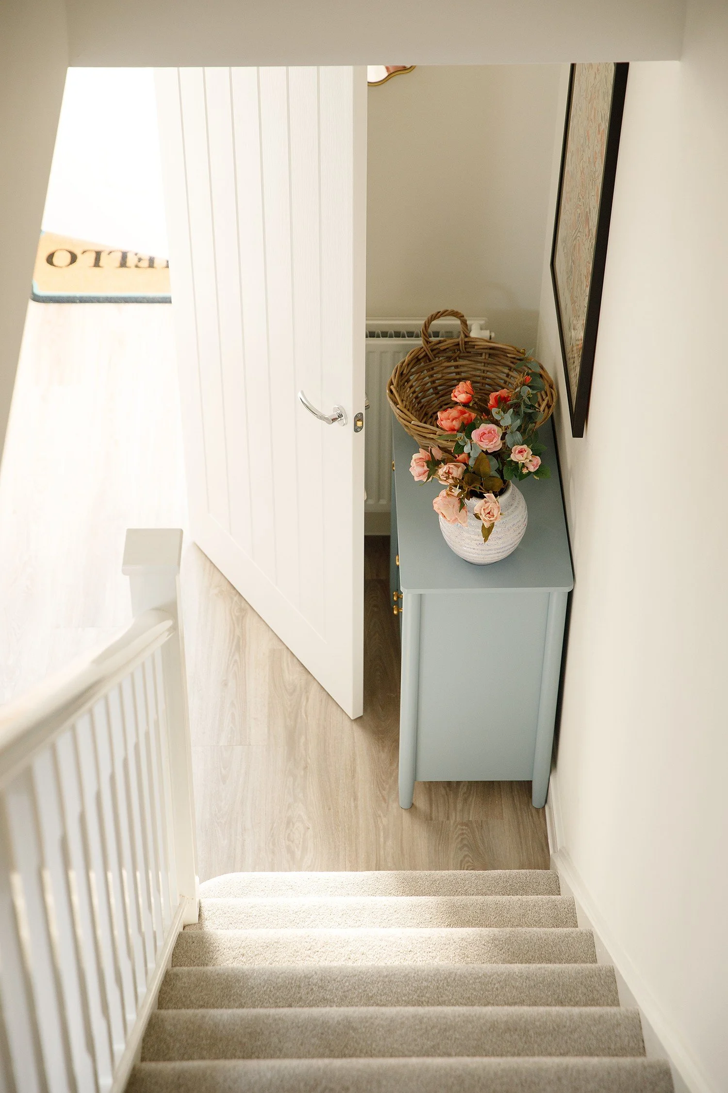 View of a staircase leading down to a decorated area with a light blue console table, a vase of pink roses, a wicker basket, and framed wall art.