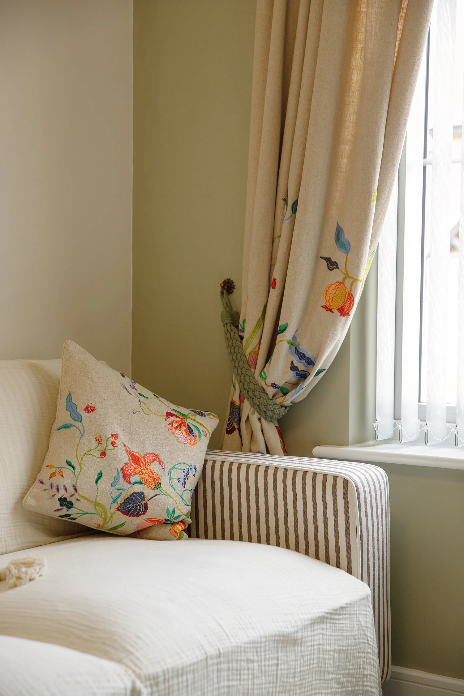A cozy bedroom corner with a bed covered in a white textured blanket, embroidered pillows with colorful floral and butterfly patterns, striped armchair, striped and floral curtains tied back, and a window allowing natural light to fill the room.