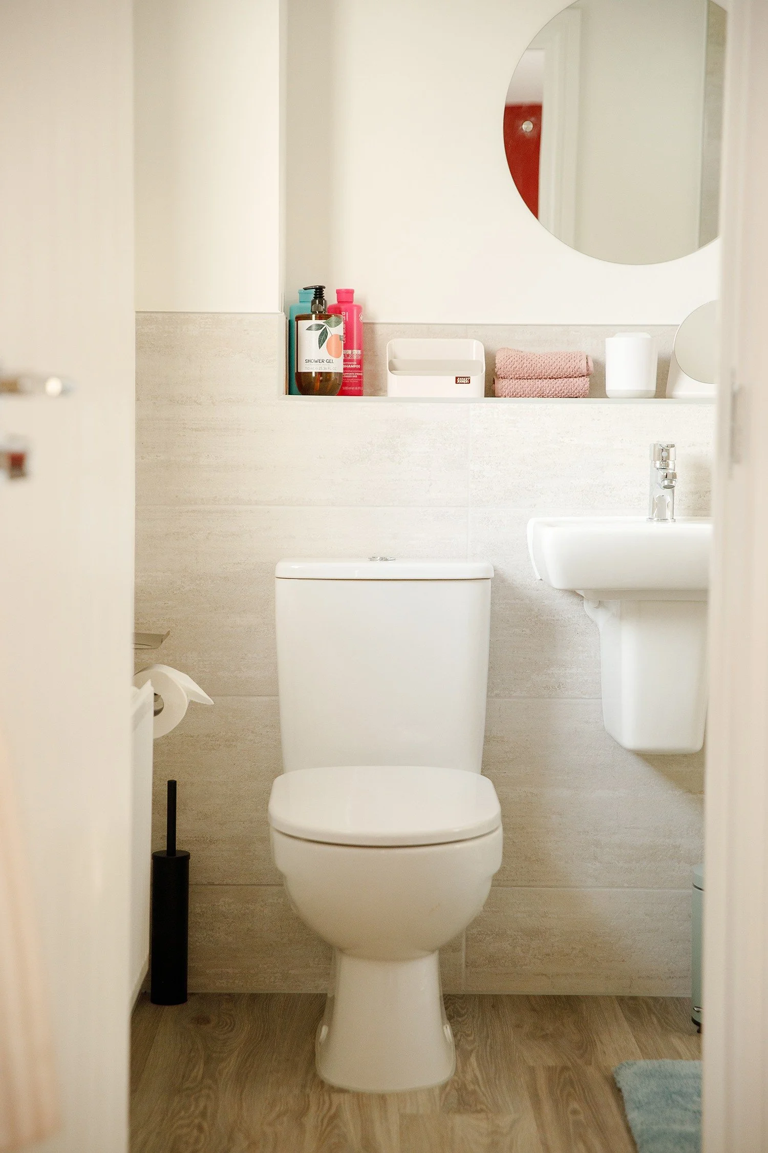 A bathroom with a toilet, small sink, mirror, toilet paper, and various toiletry bottles on a shelf.