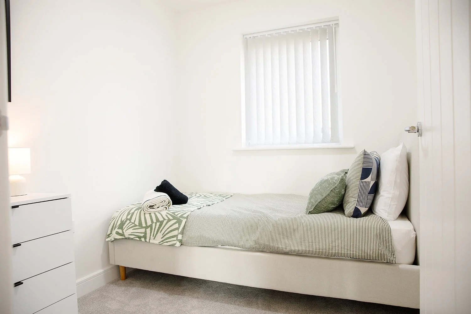 A neatly made bed with striped and patterned pillows in a bright bedroom with white walls, a window with vertical blinds, and a white nightstand with a lamp.