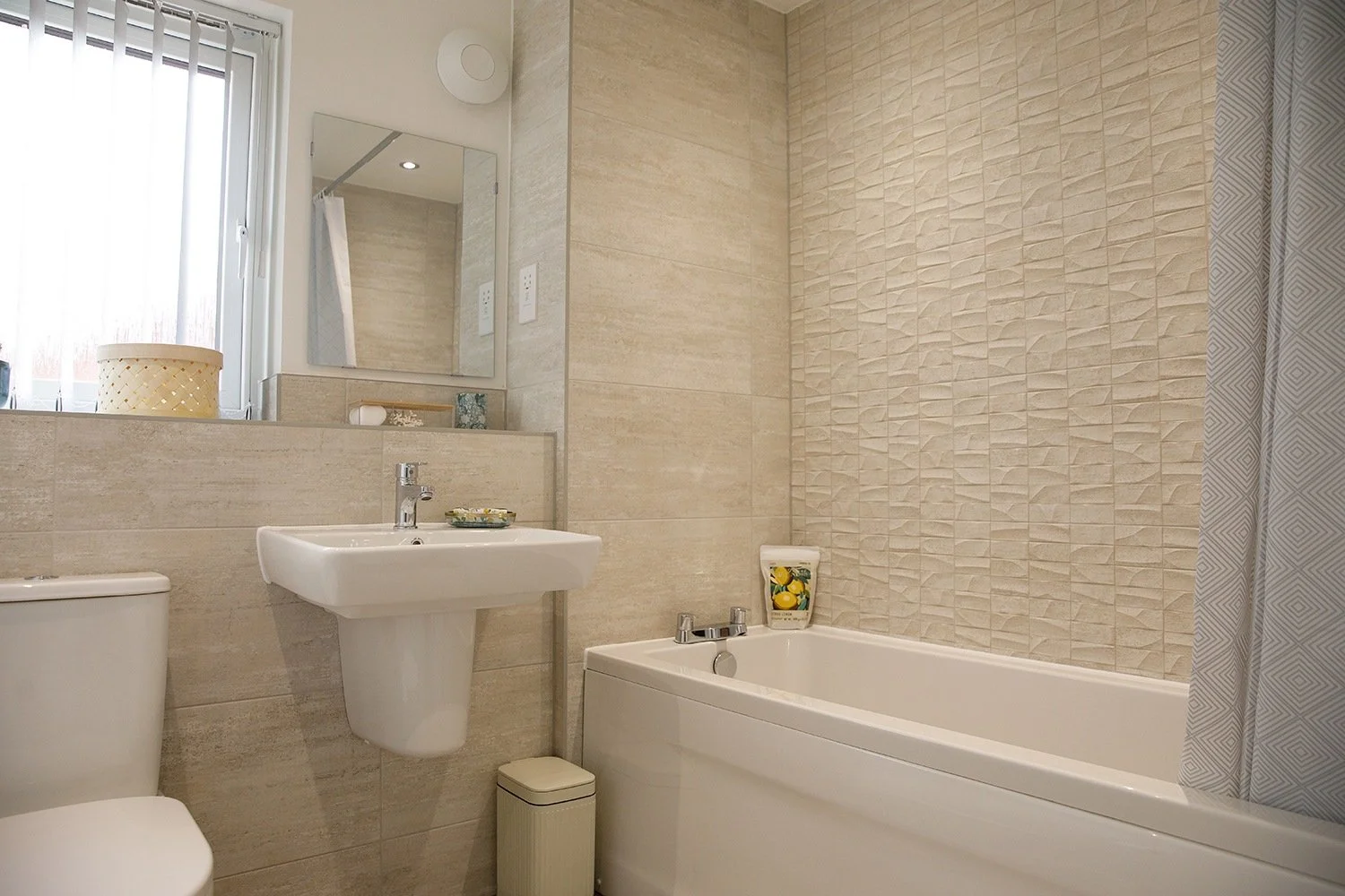 Small bathroom with beige tiles, a bathtub, a wall-mounted sink, a toilet, a mirror, and a window with curtains.
