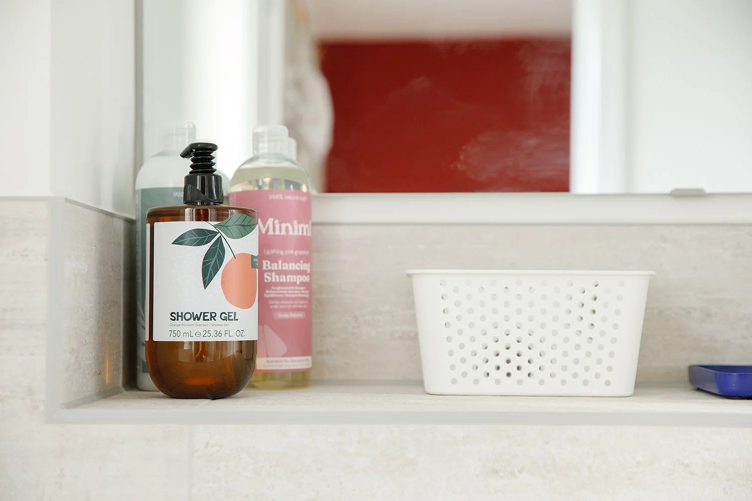 Bathroom shelf with bottles of shower gel and shampoo, an empty white perforated basket, and a blue soap dish.