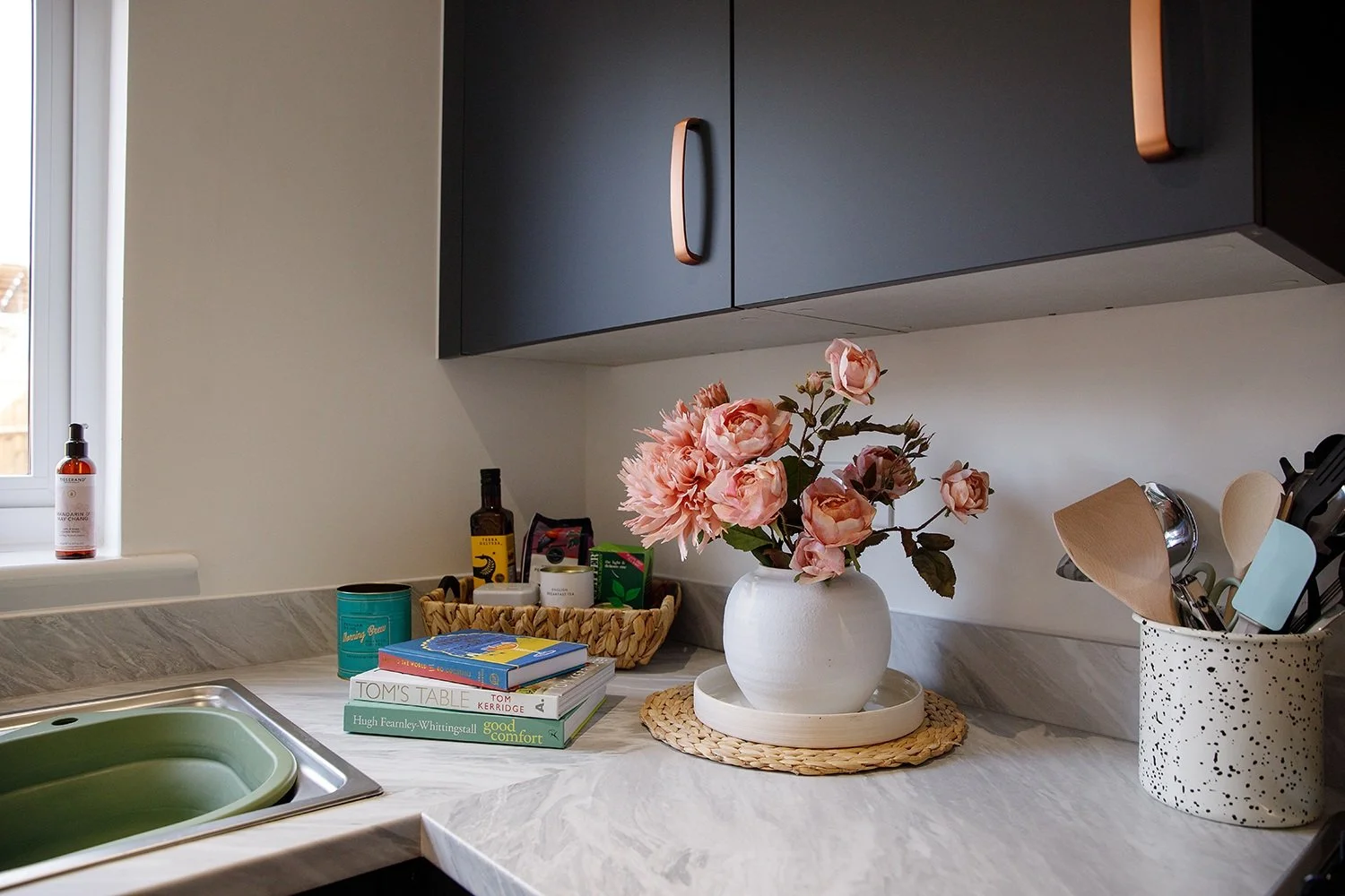 Kitchen countertop with pink flowers in a white vase, books, kitchen utensils, and various small items near a window.