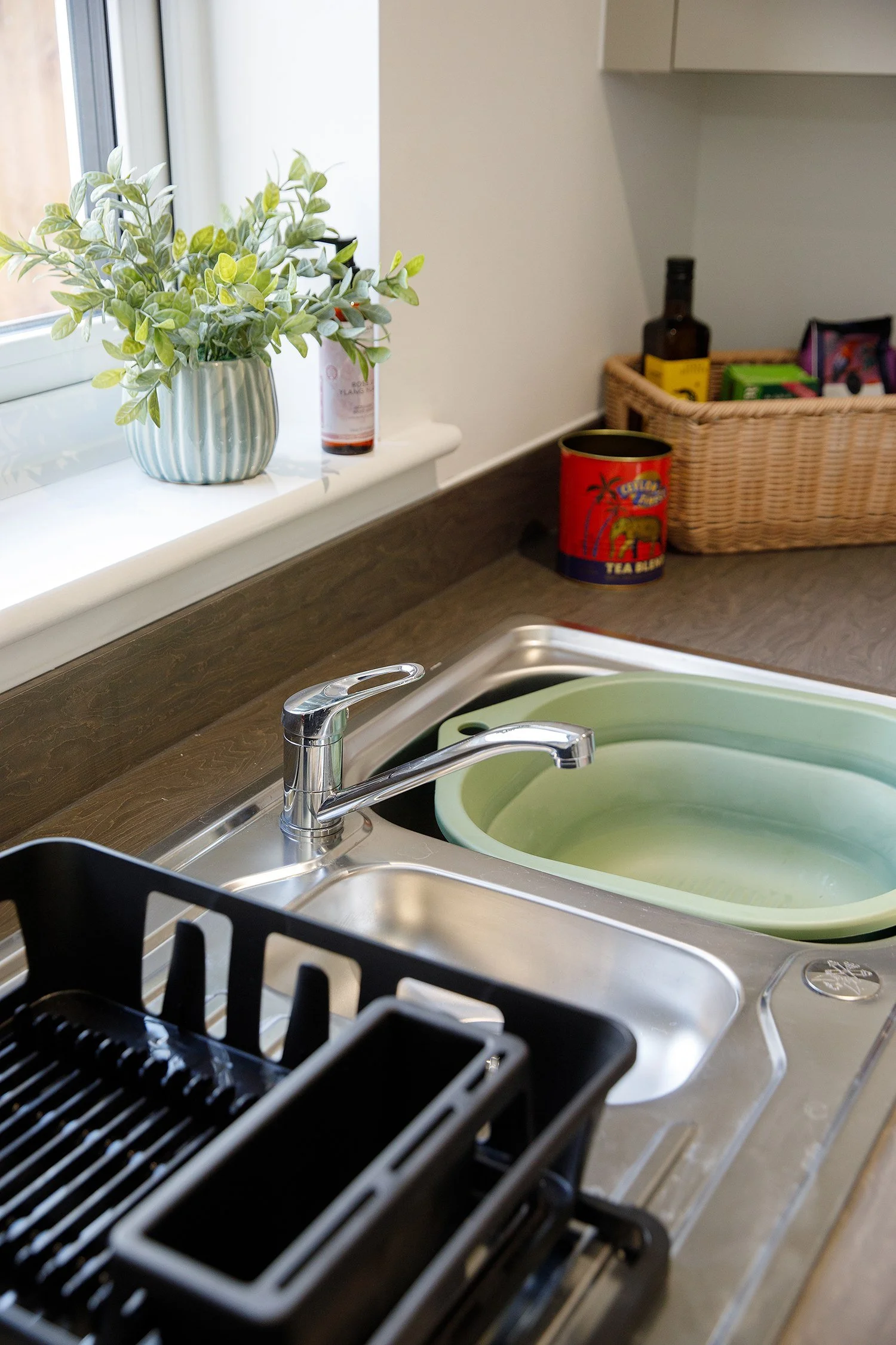 Kitchen sink with a green basin, a black dish drying rack, and a window ledge with a potted plant, a bottle, and a can of tea.