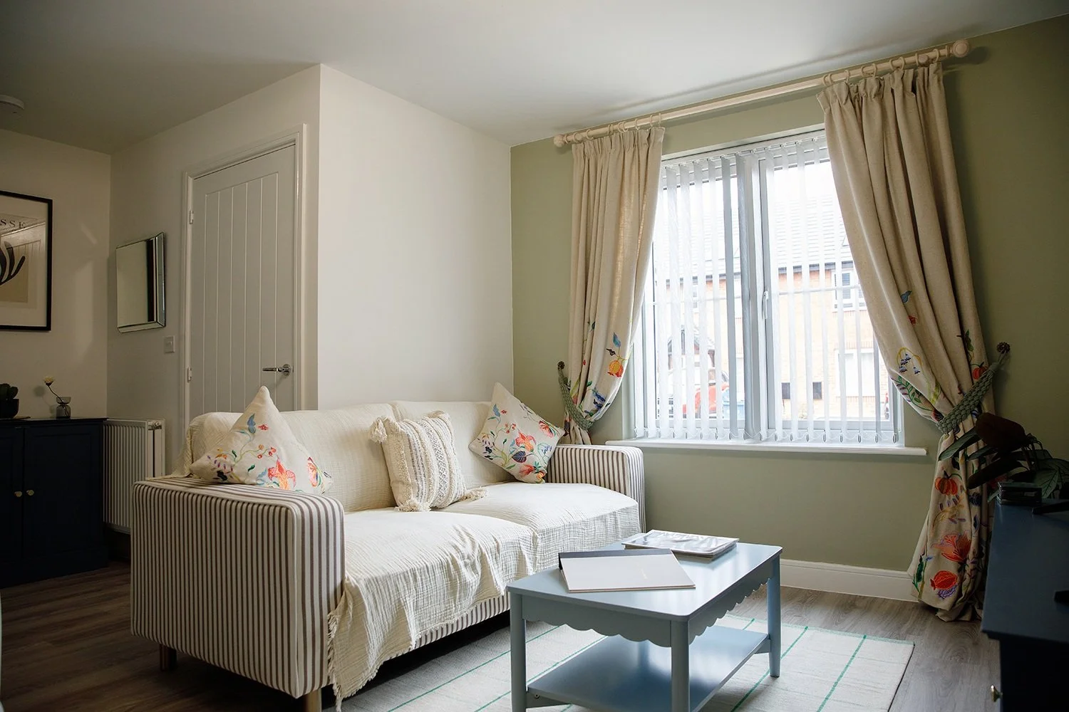 Living room with a white sofa, decorative pillows, a light blue coffee table, window with floral curtains, and a dark cabinet.