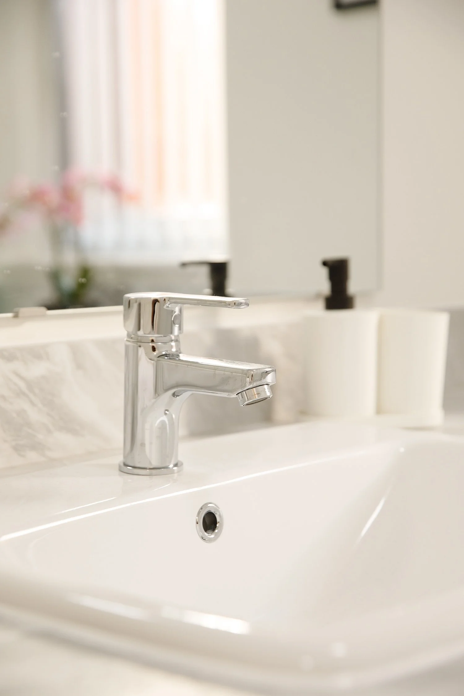 Close-up of a shiny silver bathroom faucet mounted on a white sink, with in the background a mirror reflecting a blurred view of a window with light-colored curtains and a small vase with pink flowers.