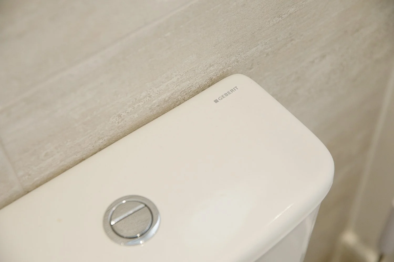 Close-up of a white Geberit toilet tank with a silver flush button, set against a beige tiled wall.