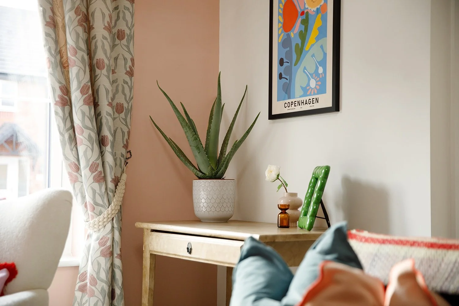 Decorative indoor scene with a cream-colored armchair, a window with floral curtains, a wooden table with potted plants, a framed colorful artwork labeled 'Copenhagen,' and a person lounging on a couch with visible legs and shoes.