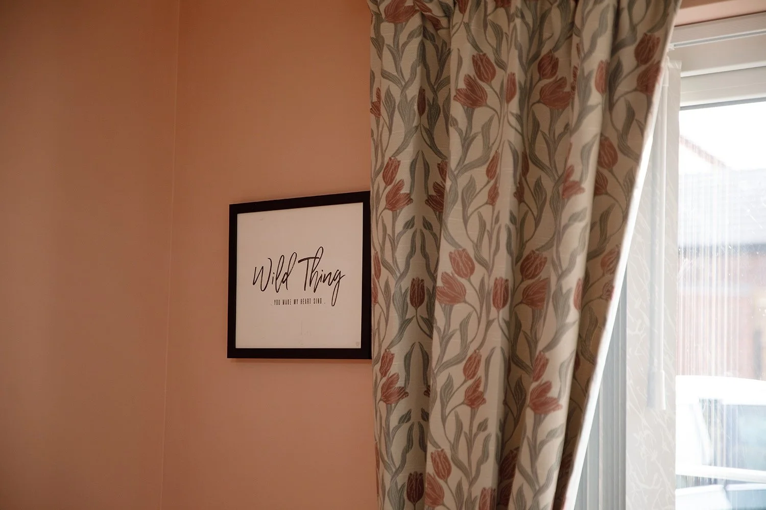 Decorative wall with framed quote, partially covered by floral patterned curtain, next to a window with sunlight.