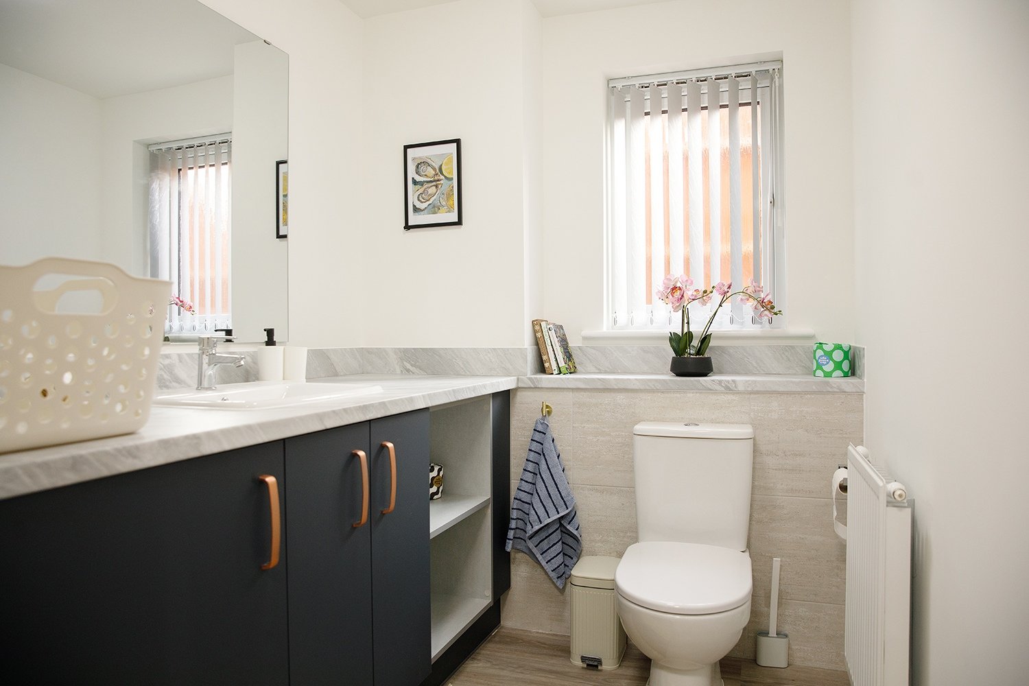 Modern bathroom with white walls, a large mirror, a blue vanity with copper handles, a white countertop, a sink with a faucet, a toilet, a window with vertical blinds, pink orchids on the windowsill, framed artwork on the wall, and a striped towel ha