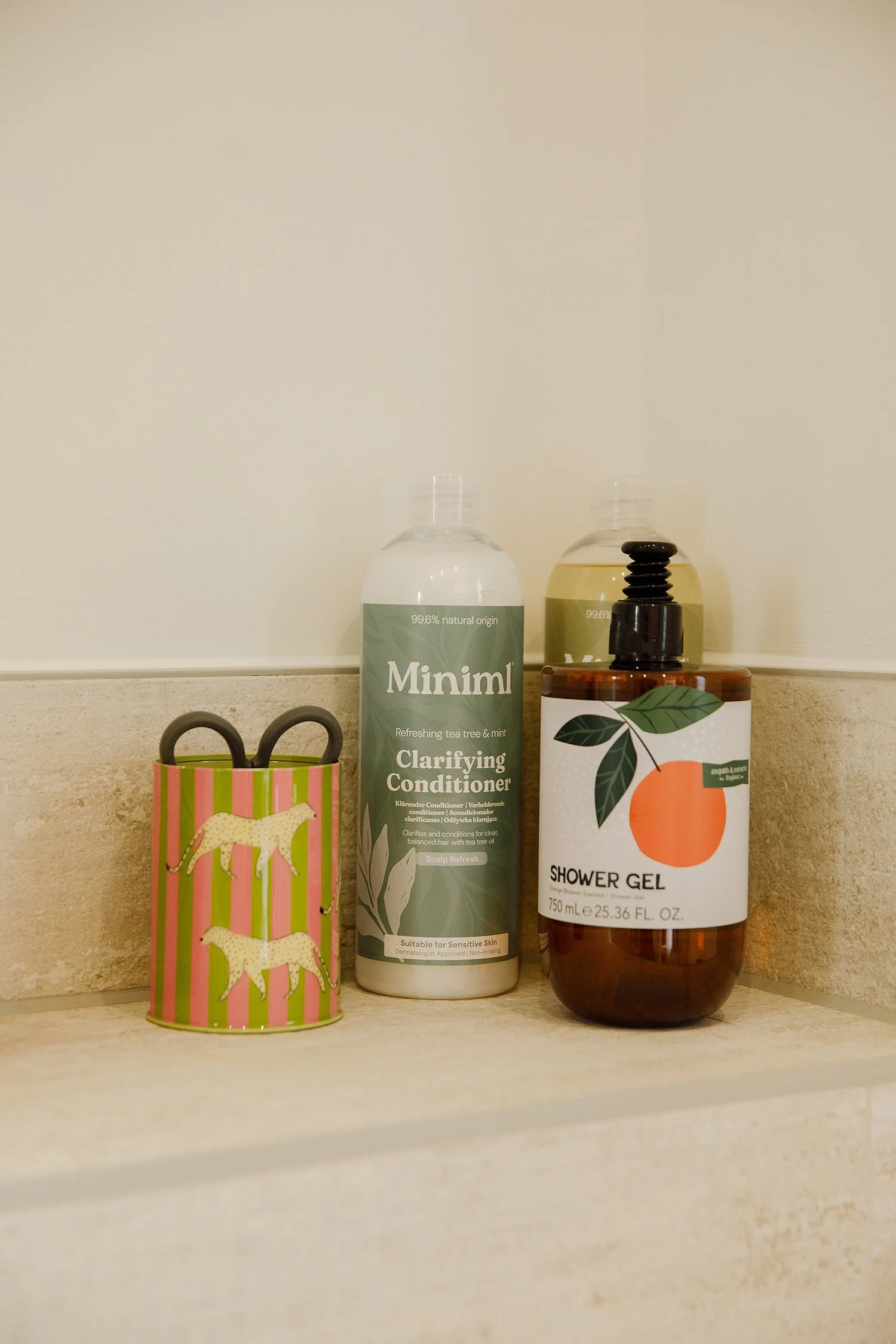 A colorful gift bag with a giraffe pattern, two bottles of hair or skincare products, and a bottle of shower gel on a beige countertop against a plain white wall.