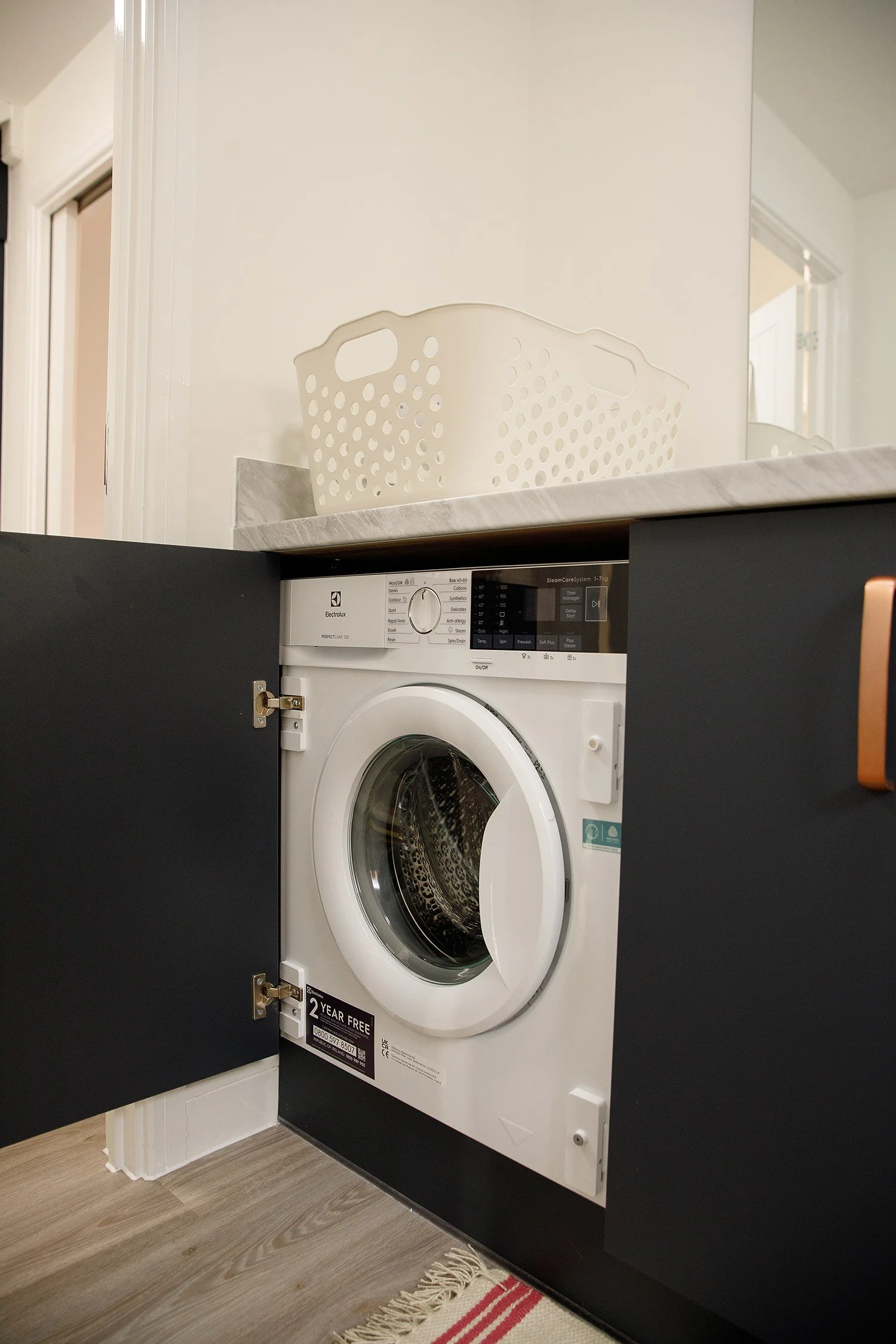 White front-loading washing machine with open black door in a laundry room with light wooden flooring and a red and white striped rug, countertop above, and a white plastic laundry basket on the countertop.