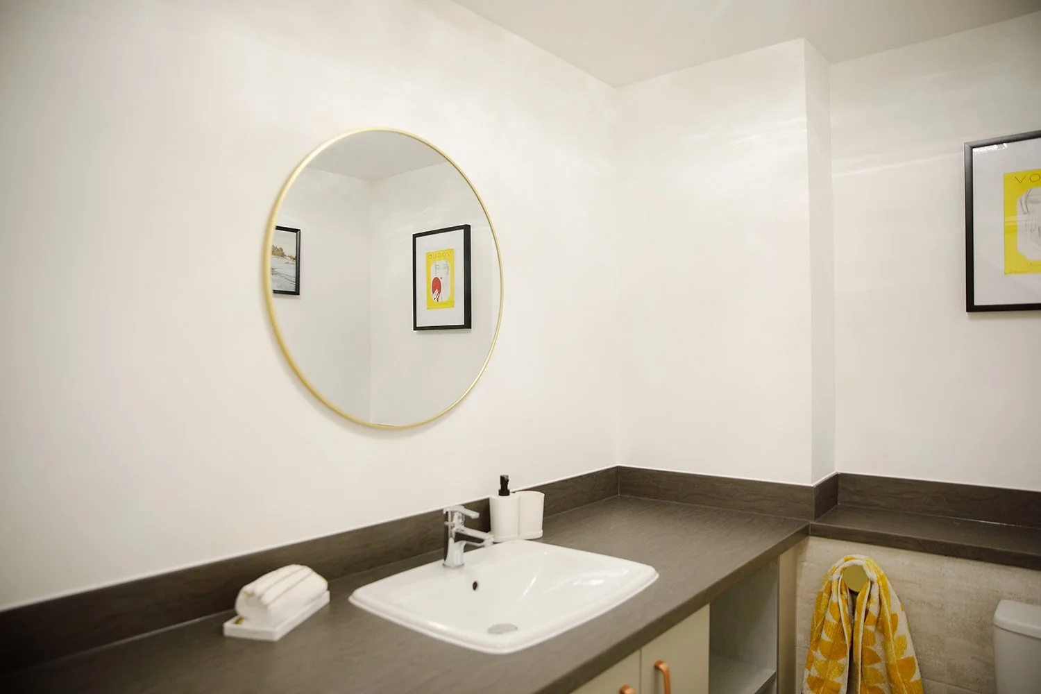 Modern bathroom with a white sink, a round mirror, framed artwork on the wall, and a yellow towel hanging on a hook.