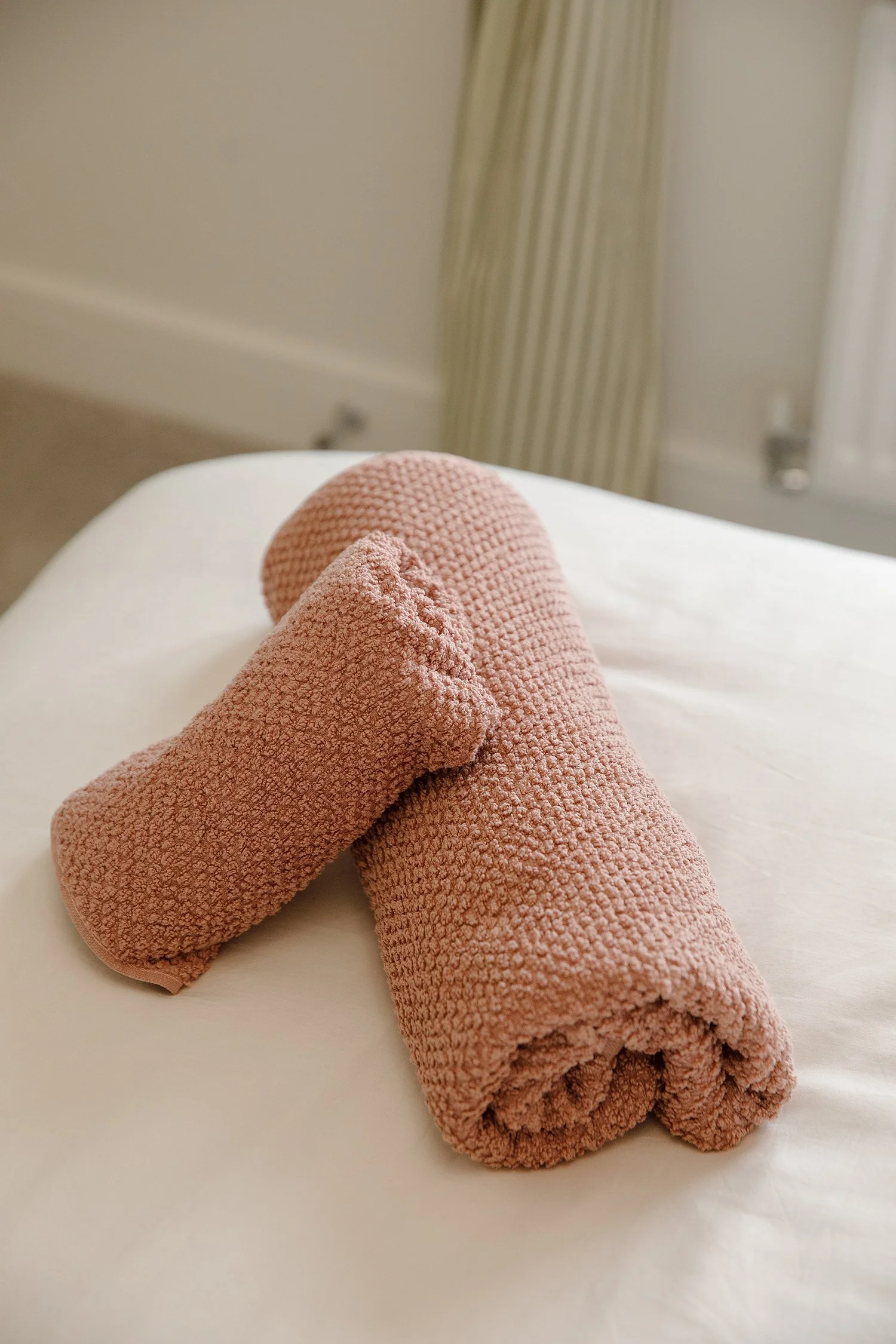 Light pink textured towel or cloth rolled up on a surface, with a similar towel partially unrolled nearby, in a room with beige walls and a radiator.