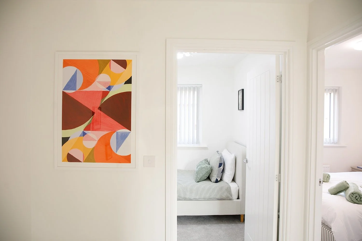 Photo of a white wall with colorful abstract art on the left and two open doorways leading to bedrooms with white walls and bedding, one with a bed and pillows, the other with rolled towels.