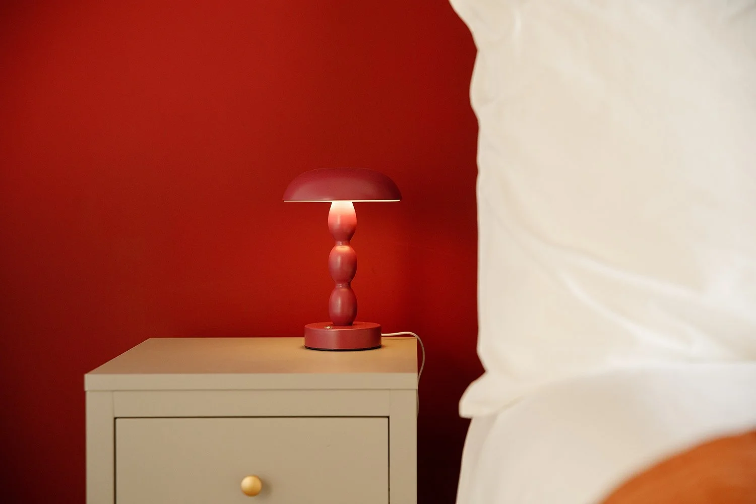 A pink table lamp with a bulbous base and a pink lampshade is placed on a white nightstand against a dark red wall, partially obscured by a white pillow in the foreground.