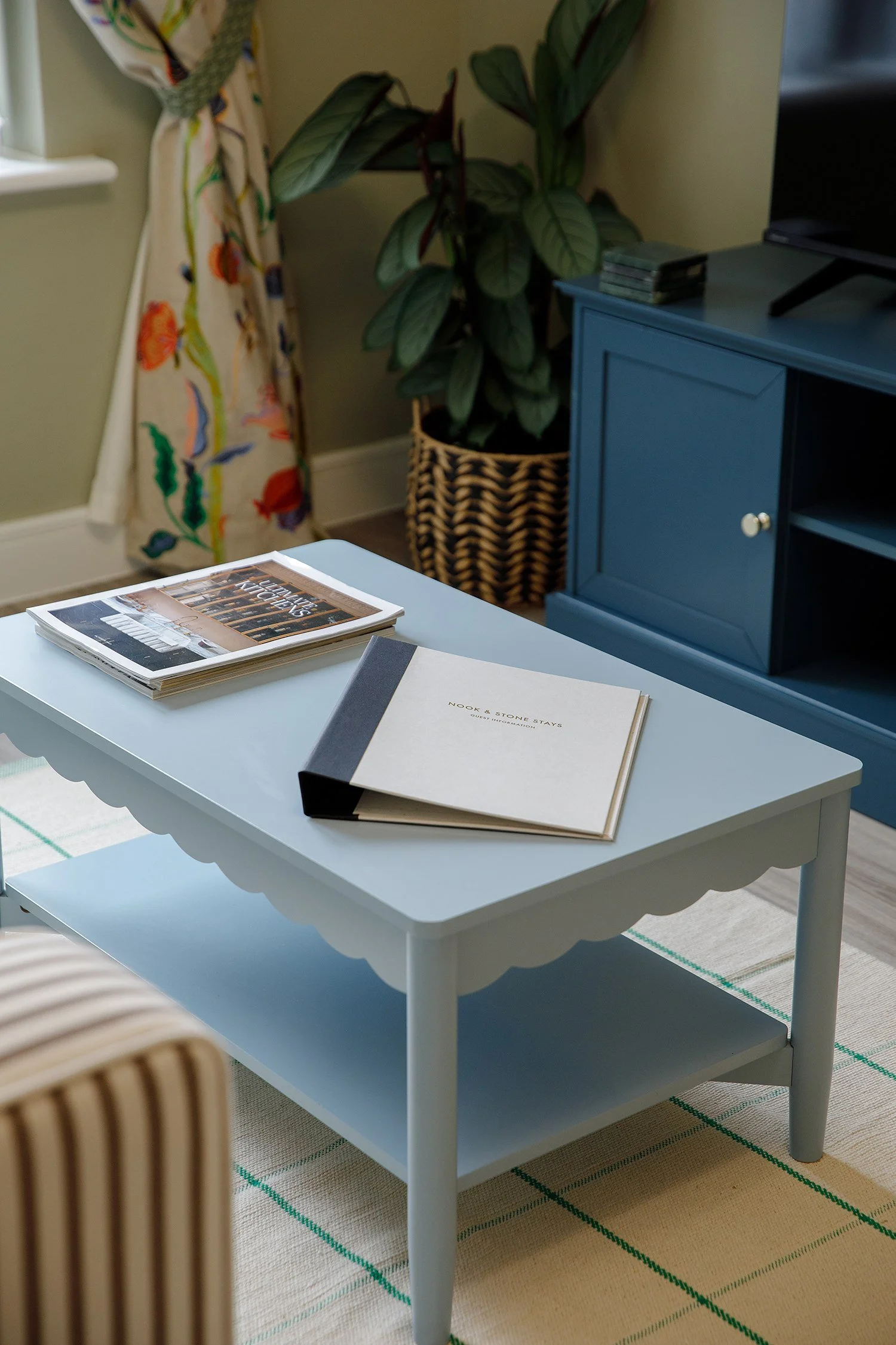 A white coffee table with a lower shelf, holding magazines and a guest book, located in a room with a framed rug on a beige carpet, a plant in a wicker basket, a blue cabinet, and a colorful curtain.