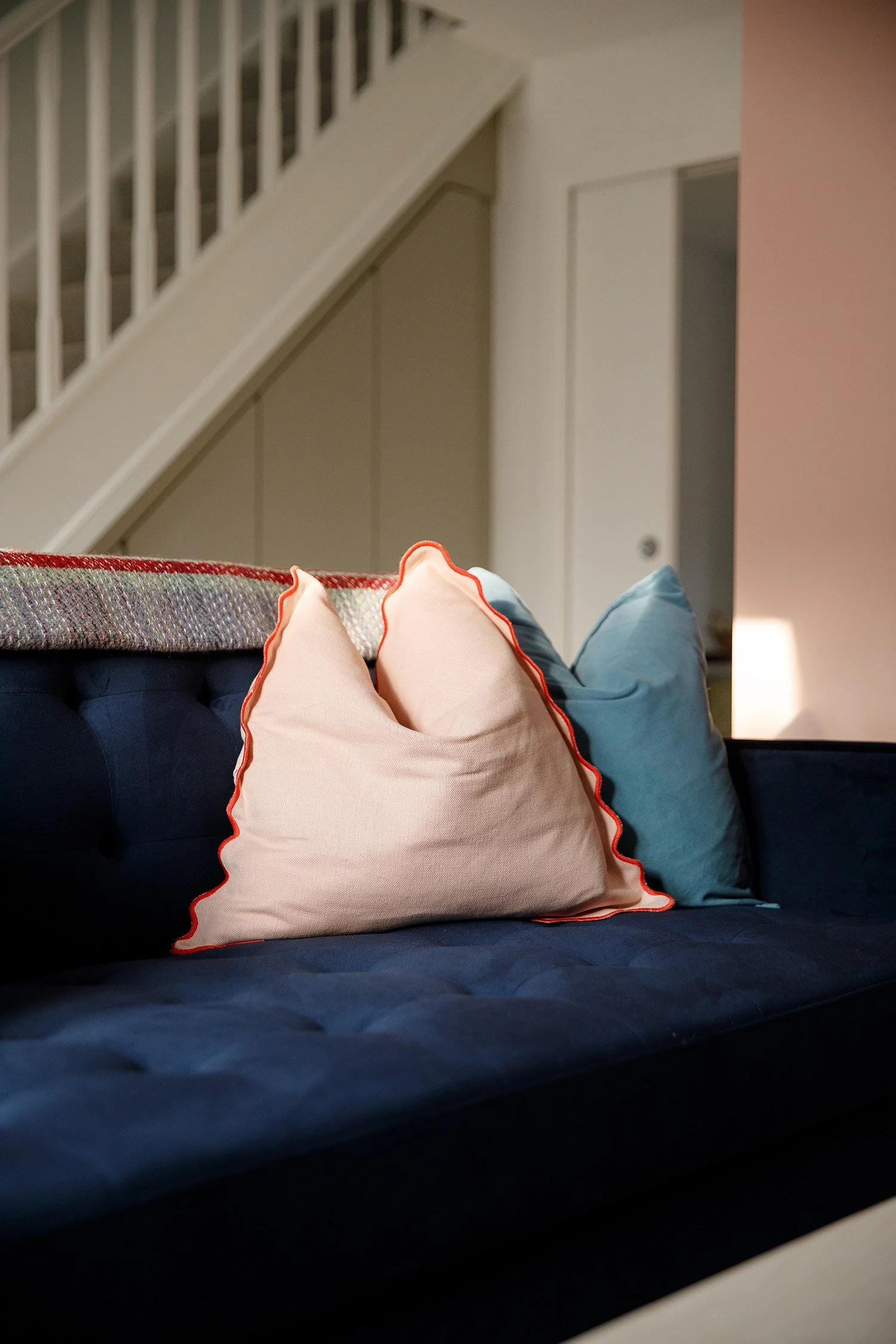 A living room scene with a navy sofa holding several decorative pillows, including a peach pillow with orange trim, a blue pillow, and a white pillow. In the background, a staircase with white railing and beige wall panels are visible.
