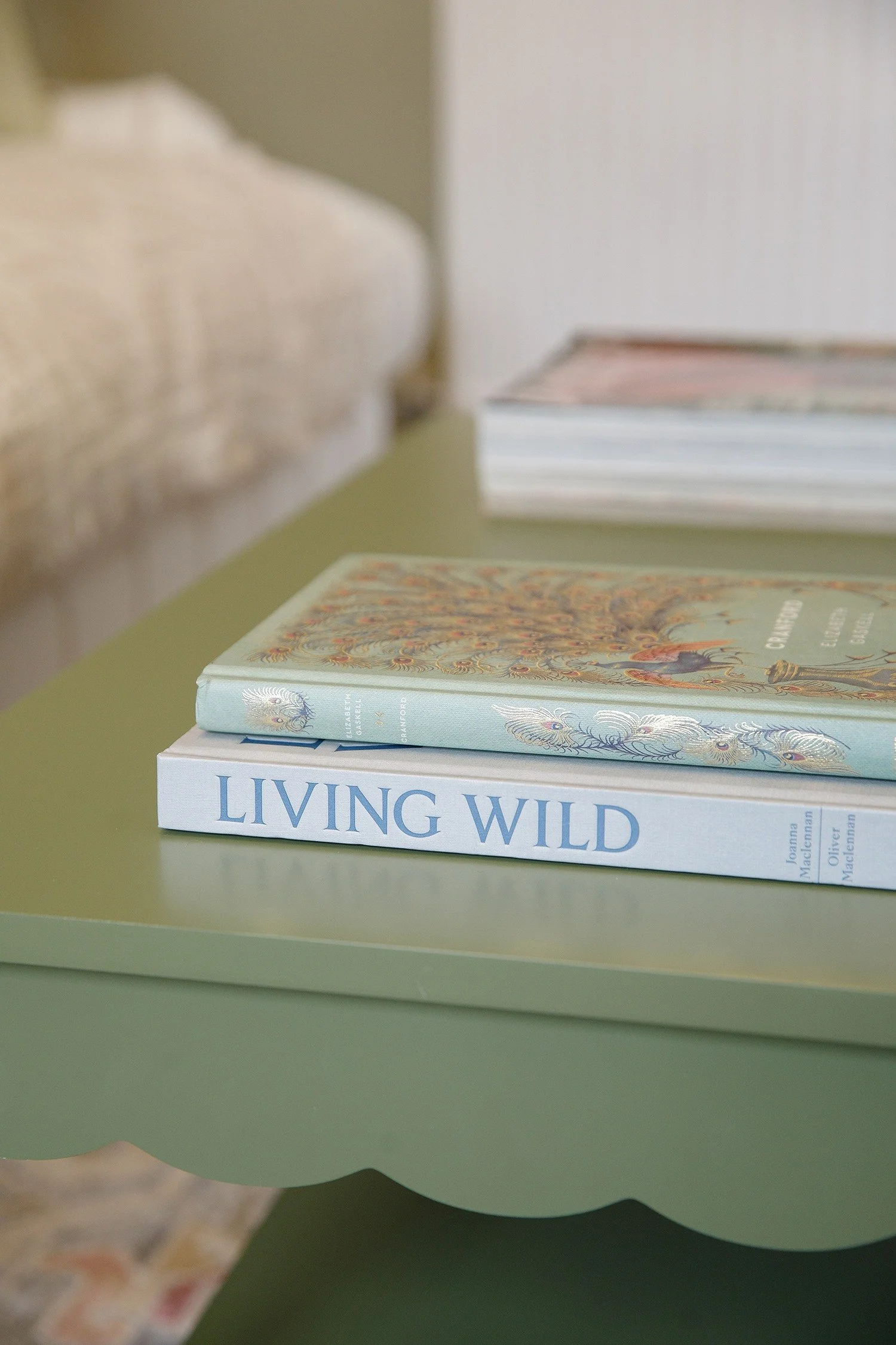 Stack of books on a green table with the title "Living Wild" visible, and a second book with a colorful cover. The background shows a blurry partial view of the table's edge and a patterned rug.