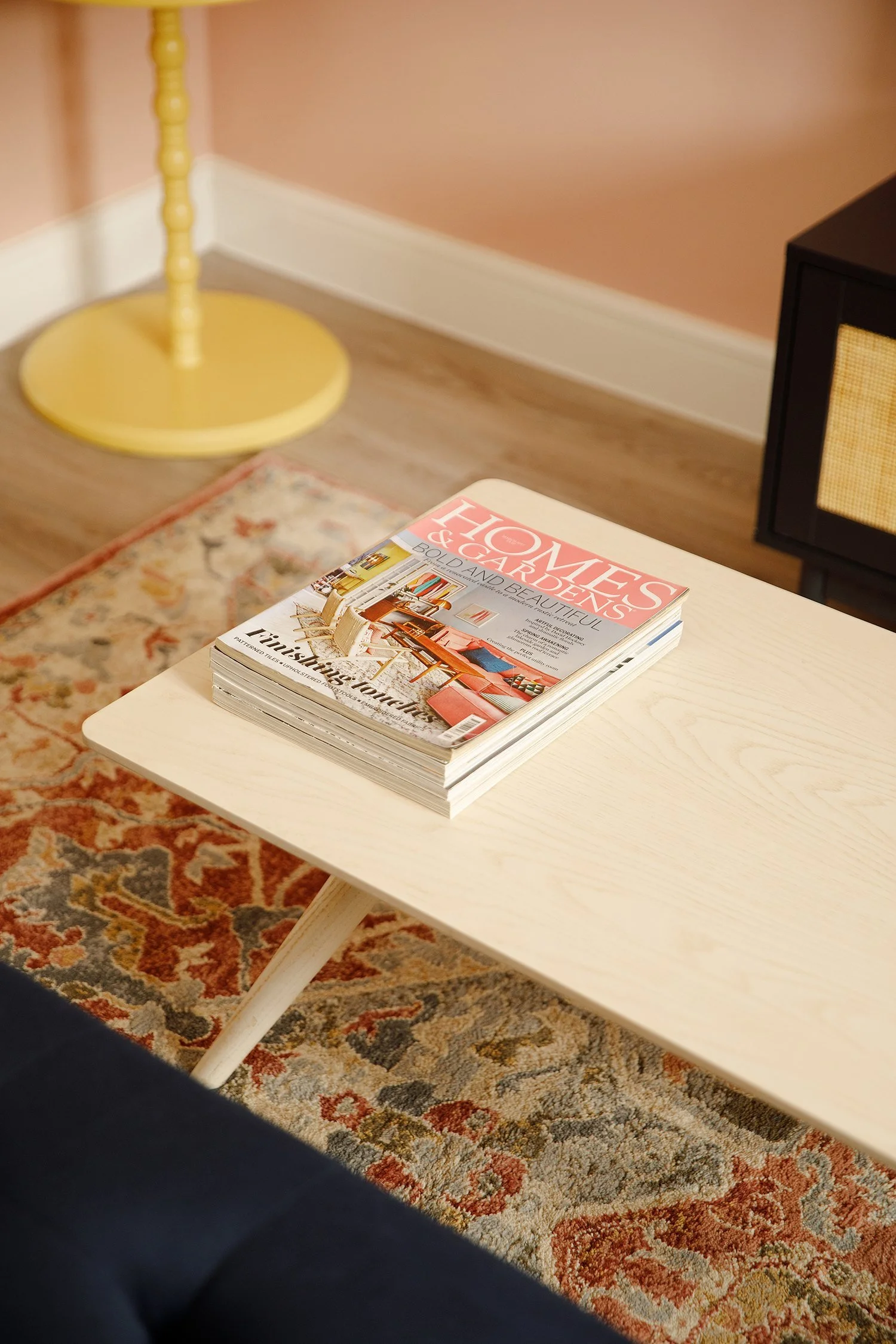 A stack of magazines on a white table with a floral rug underneath, a yellow lamp in the corner, against a peach-colored wall and a black cabinet.