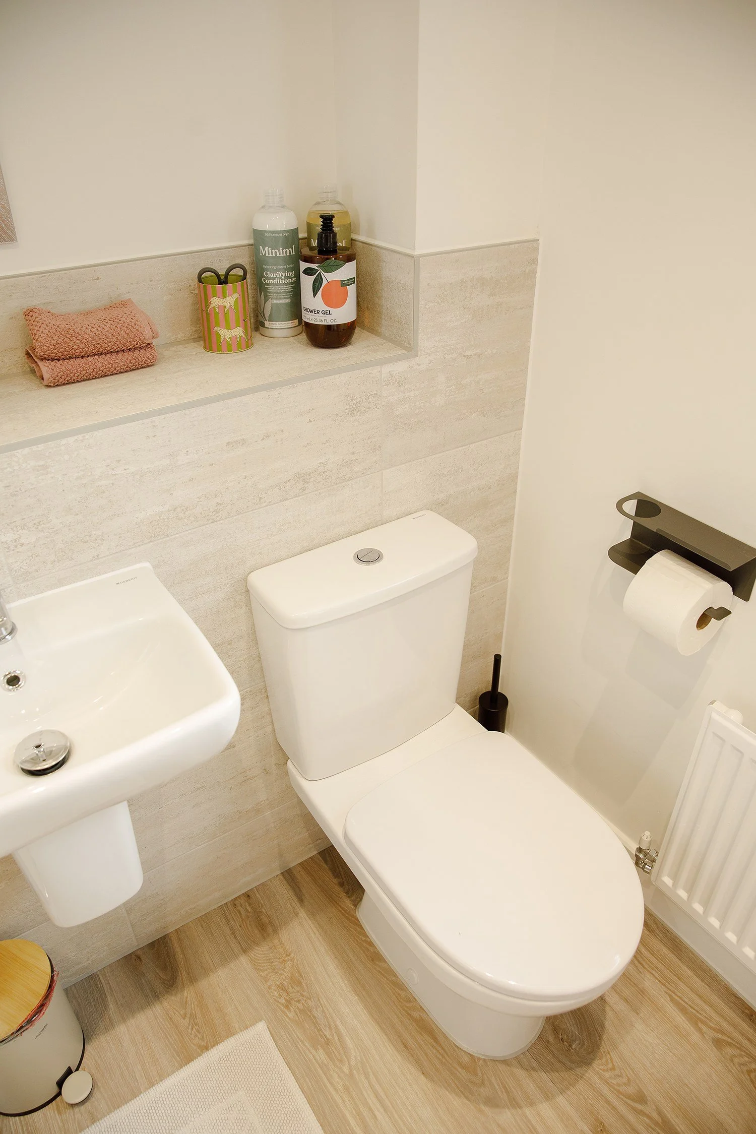 A bathroom with a white toilet, white sink, and a shelf with toiletries including shampoo, conditioner, and shower gel. There is a pink towel, a small yellow trash can, and a toilet paper holder with toilet paper.