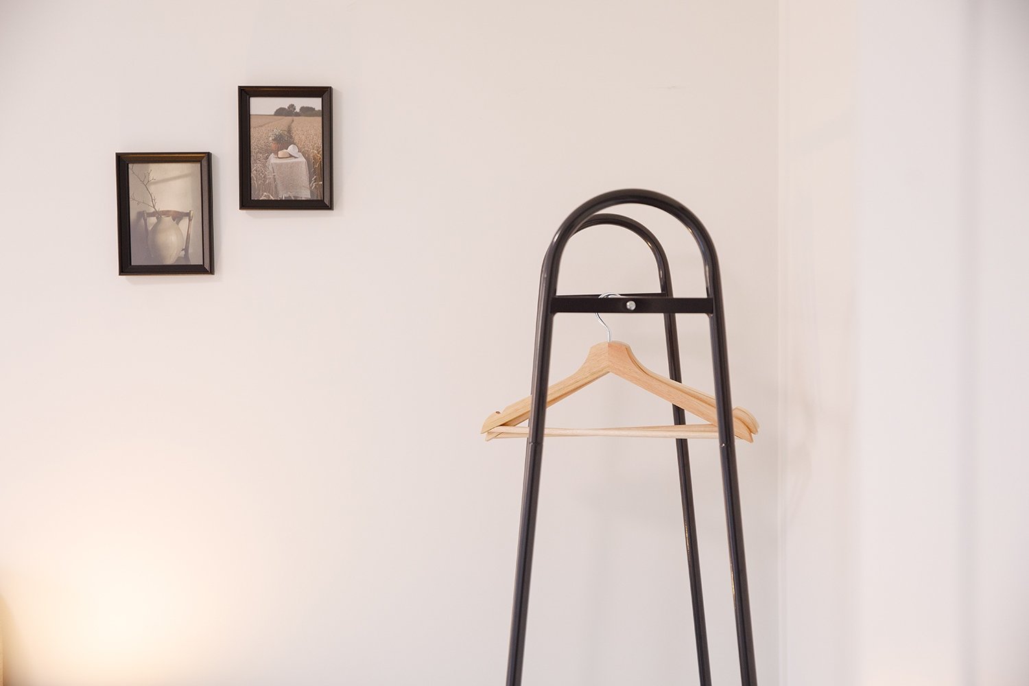 A black metal clothing rack with three legs and a wooden hanger hanging on it, positioned against a plain off-white wall. Two framed pictures are hanging on the wall, one showing a vase and the other a field with a hat on a table.