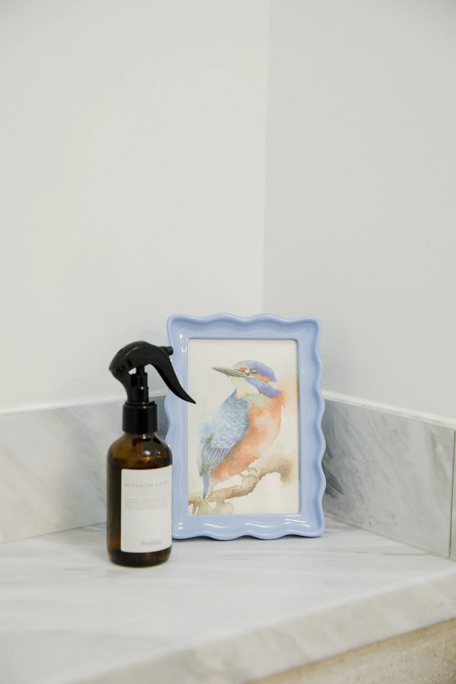 A spray bottle of cleaning solution in front of a framed watercolor painting of a colorful bird, placed on a marble countertop with a plain white wall background.
