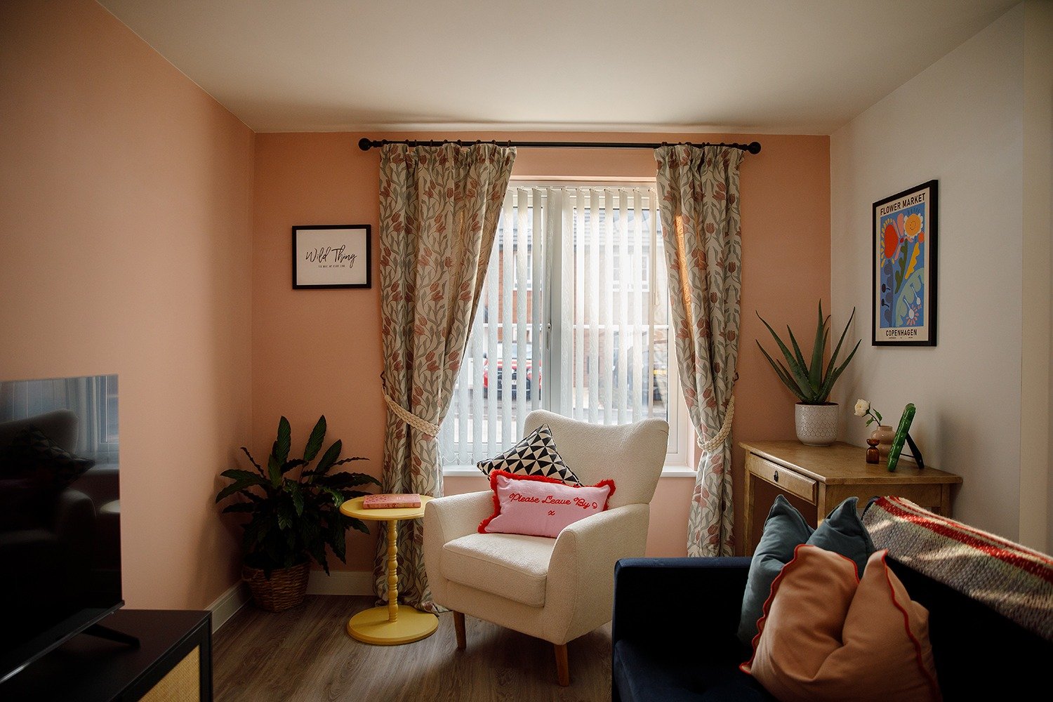 Living room with peach and white walls, a window with curtains, a white armchair with decorative pillows, a wooden side table, potted plants, and framed artwork on the walls.