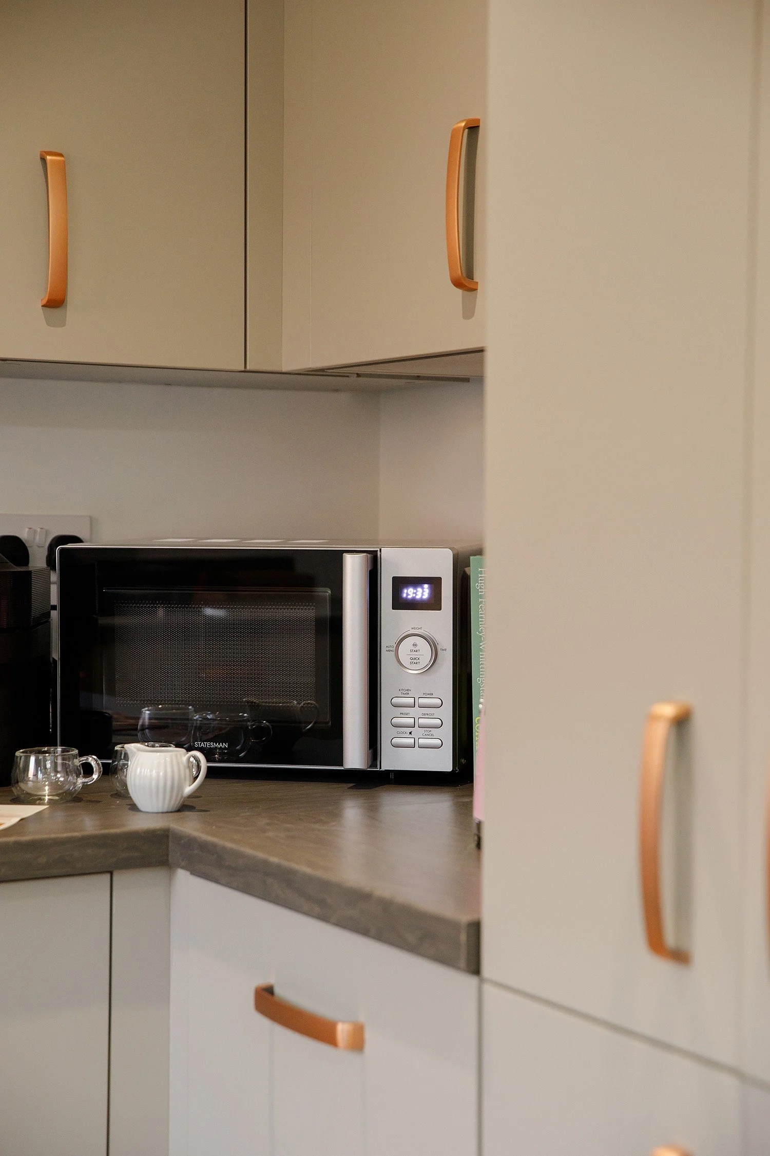 Kitchen cabinets with orange handles, microwave oven on a countertop, small glass teapots, and a creamer pitcher on a kitchen counter.