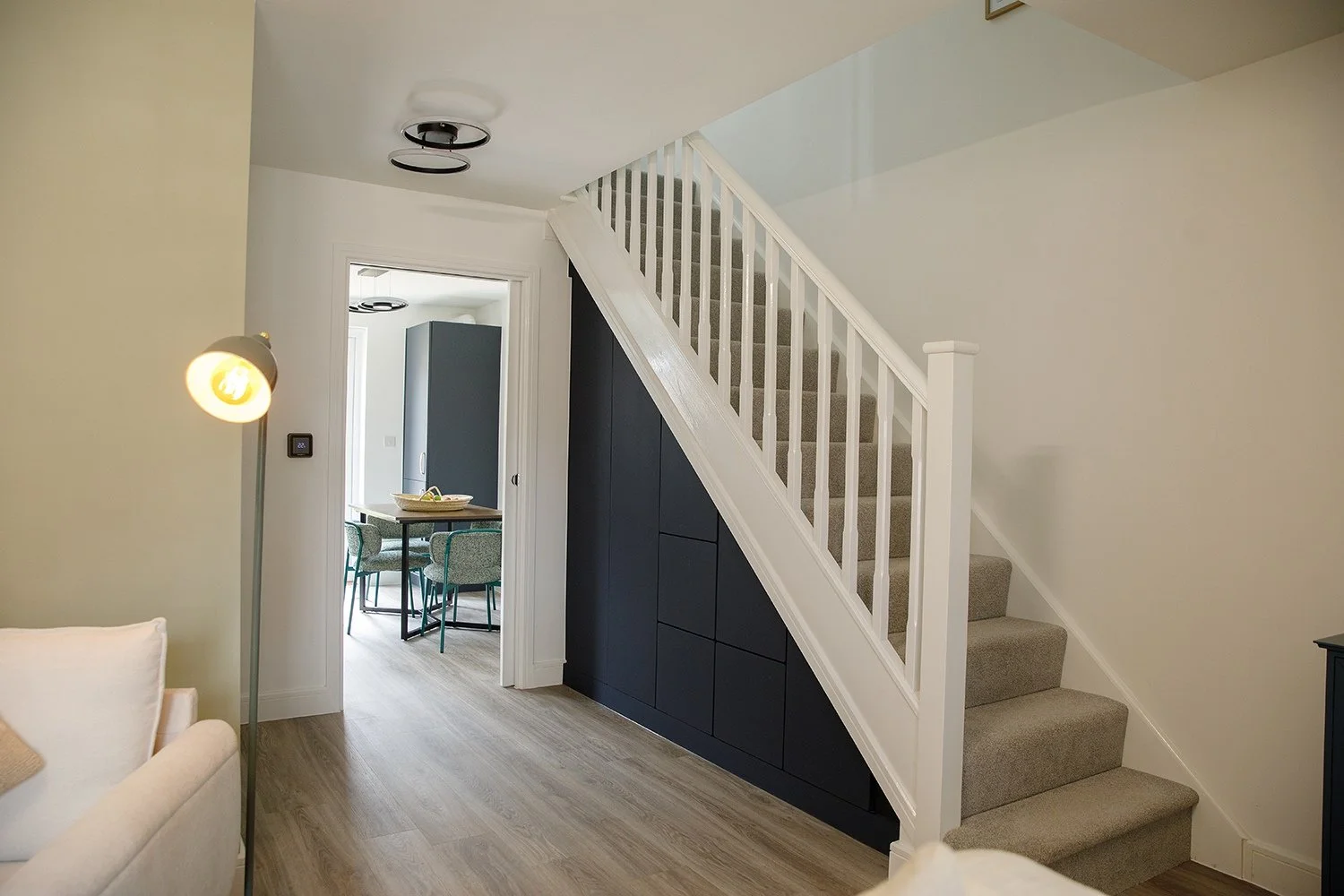 Interior view of a modern home with a staircase, black storage cabinetry beneath it, a doorway leading to a dining area with a table and chairs, and contemporary lighting fixtures.