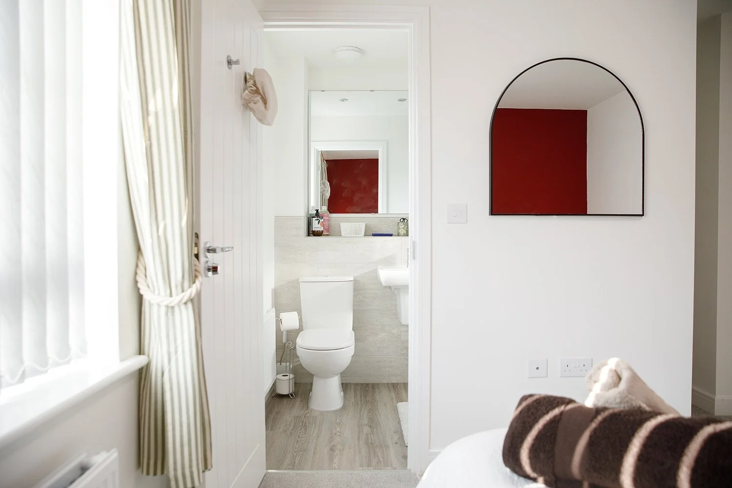 View of a white bathroom with toilet, mirror, and sink, seen from an adjoining room with a white wall, that has a round mirror with black frame, red wall reflection in the mirror, and a window with curtain.