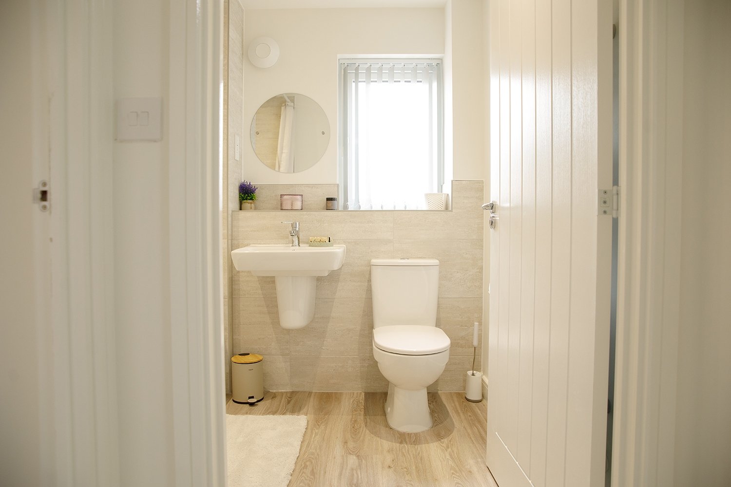 A small, clean bathroom with a white toilet, a white sink, a mirror above the sink, a small window with vertical blinds, a beige tiled wall, and hardwood flooring. Decor includes a potted plant, a soap dish, and a trash can.